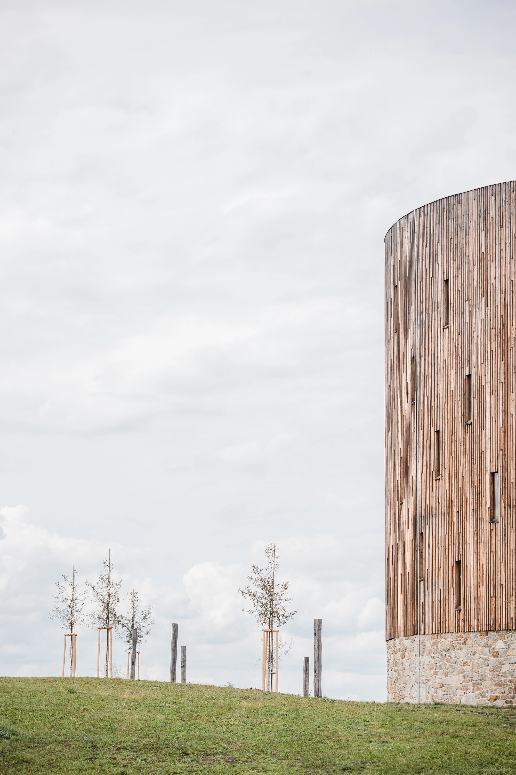 Our Lady of Sorrows Chapel by RCNKSK — A cylindrical timber-clad tower dominates the right frame, its vertical wood boarding interrupted by narrow rectangular window openings that punctuate the...