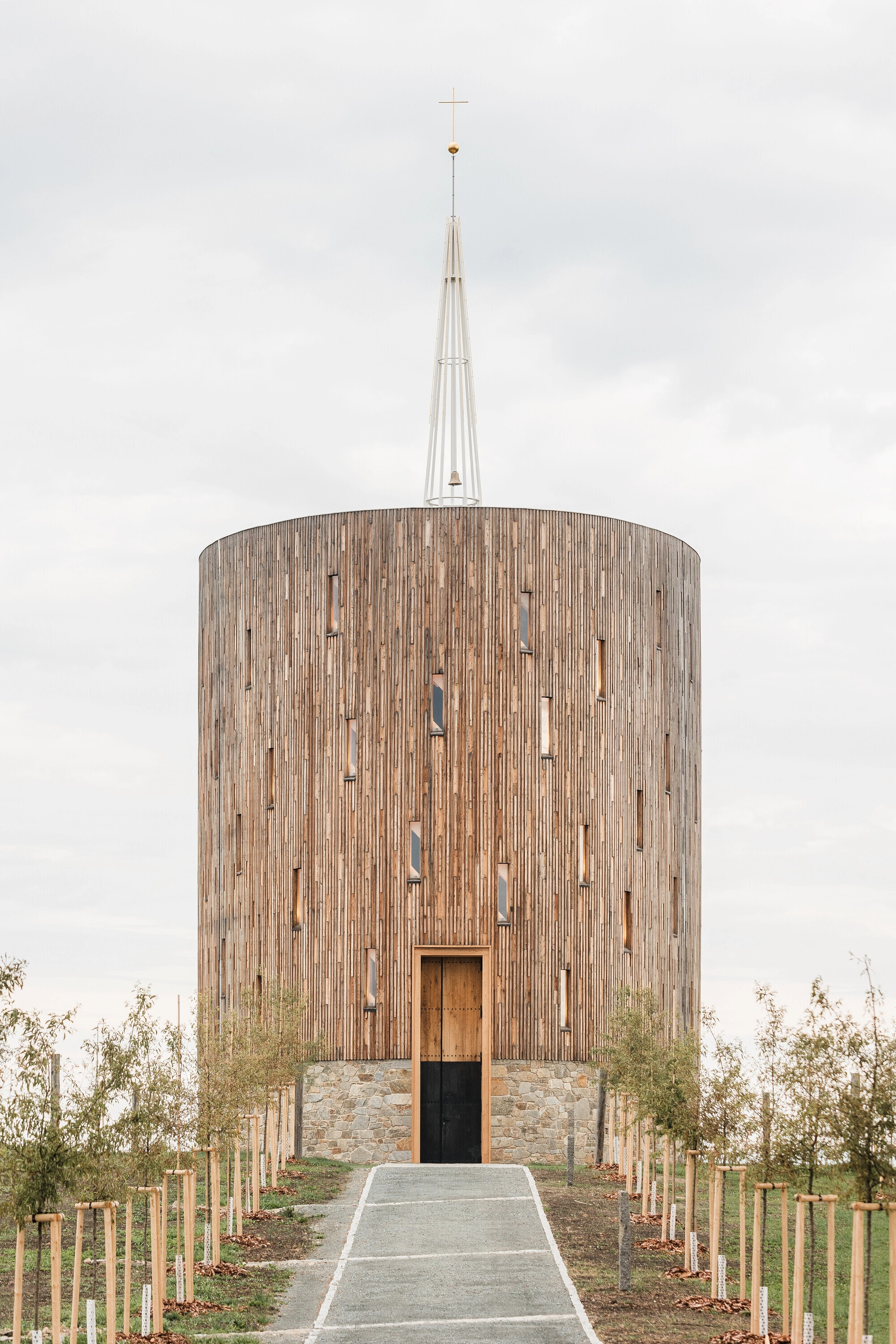 Our Lady of Sorrows Chapel by RCNKSK — Frontal view of the cylindrical timber chapel ascending symmetrically toward a slender spire topped with a brass cross.