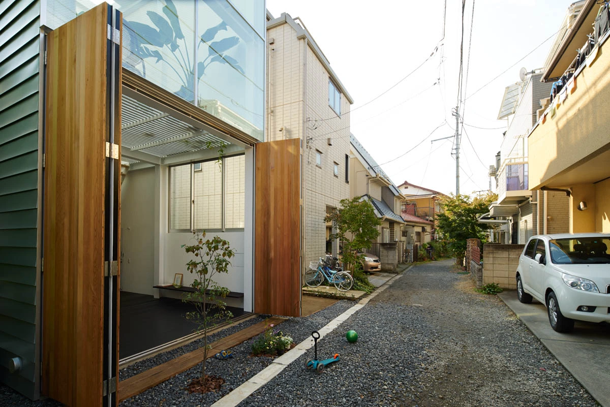 House in Kawasaki by Taichi Mitsuya & Associates — The timber-framed entrance dominates this street-level view, its warm wood cladding articulated by large folding doors that open the interior to the gravel...
