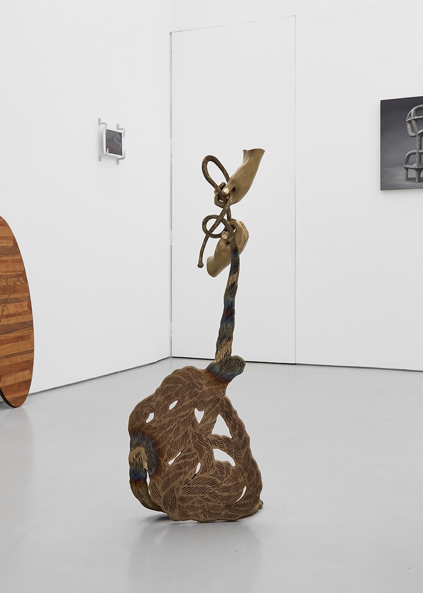Morphogenesis at Xxijra Hii — A towering bronze sculptural form dominates the gallery floor, its base a dense, organic mass with pierced negative space, rising to a slender stem that...