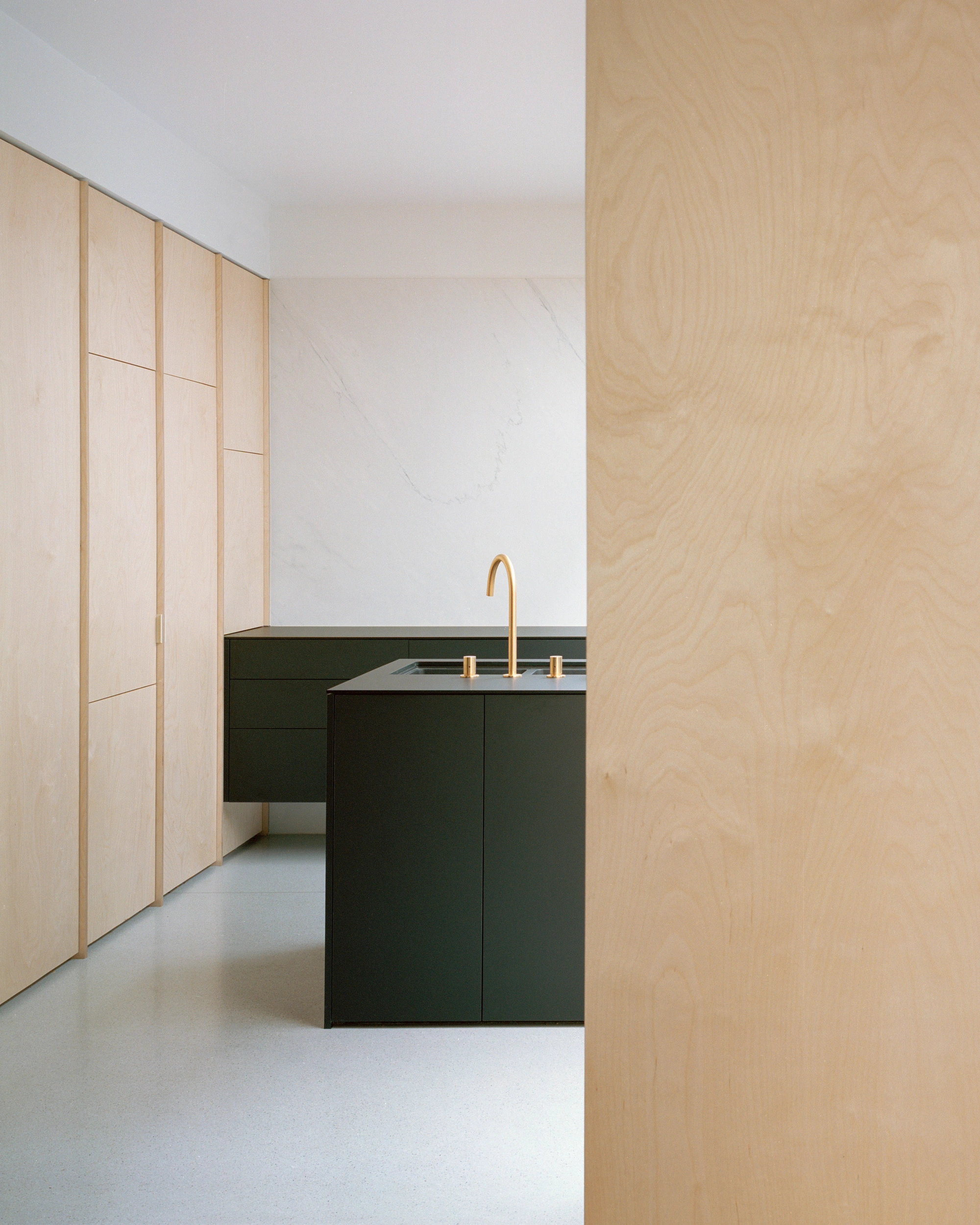 House K by Kart Projects — A minimalist kitchen intervention featuring a dark-stained timber island with brass fixtures positioned within a light timber-lined corridor.