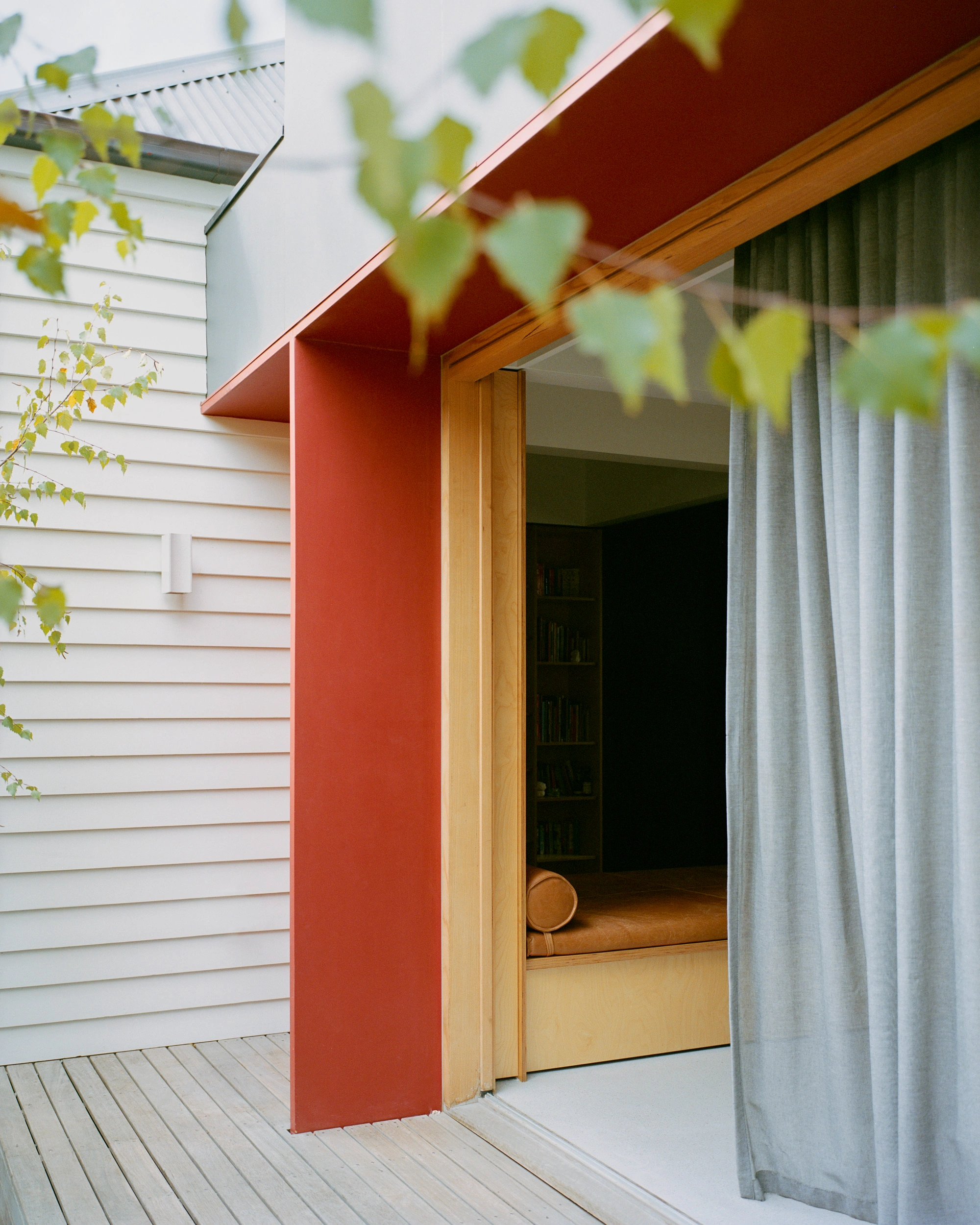 House K by Kart Projects — A lean timber-framed extension pivots open onto a pale timber deck, its deep red-orange rendered wall creating bold contrast against white horizontal...