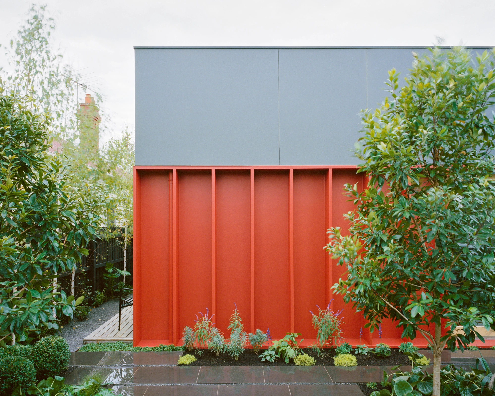 House K by Kart Projects — The garden facade of House K presents a bold chromatic strategy, with a rendered wall in crimson-red divided by vertical ribbed panels that rise from a dark...