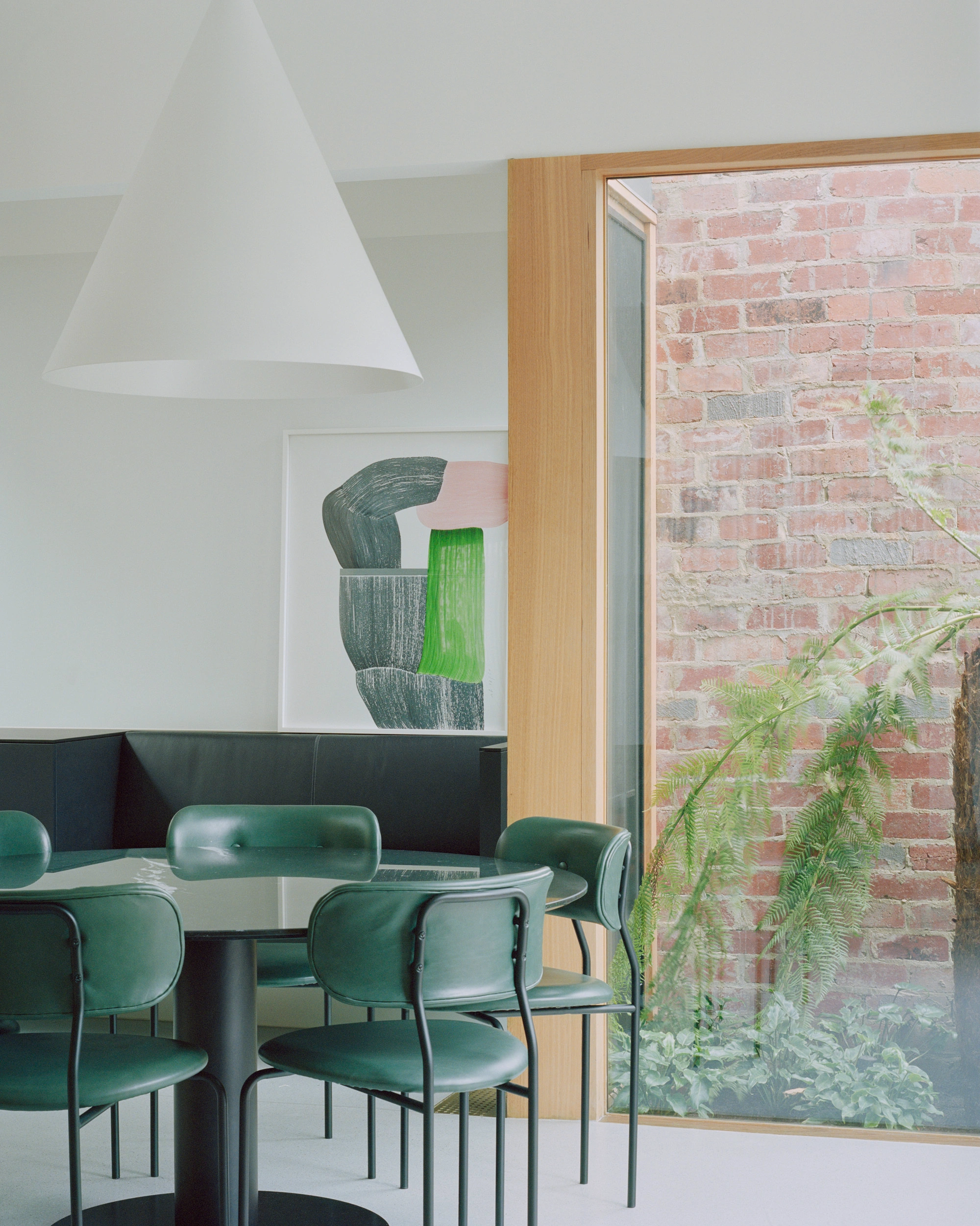 House K by Kart Projects — A dining nook at House K features green leather chairs arranged around a circular black table beneath a conical white pendant light.