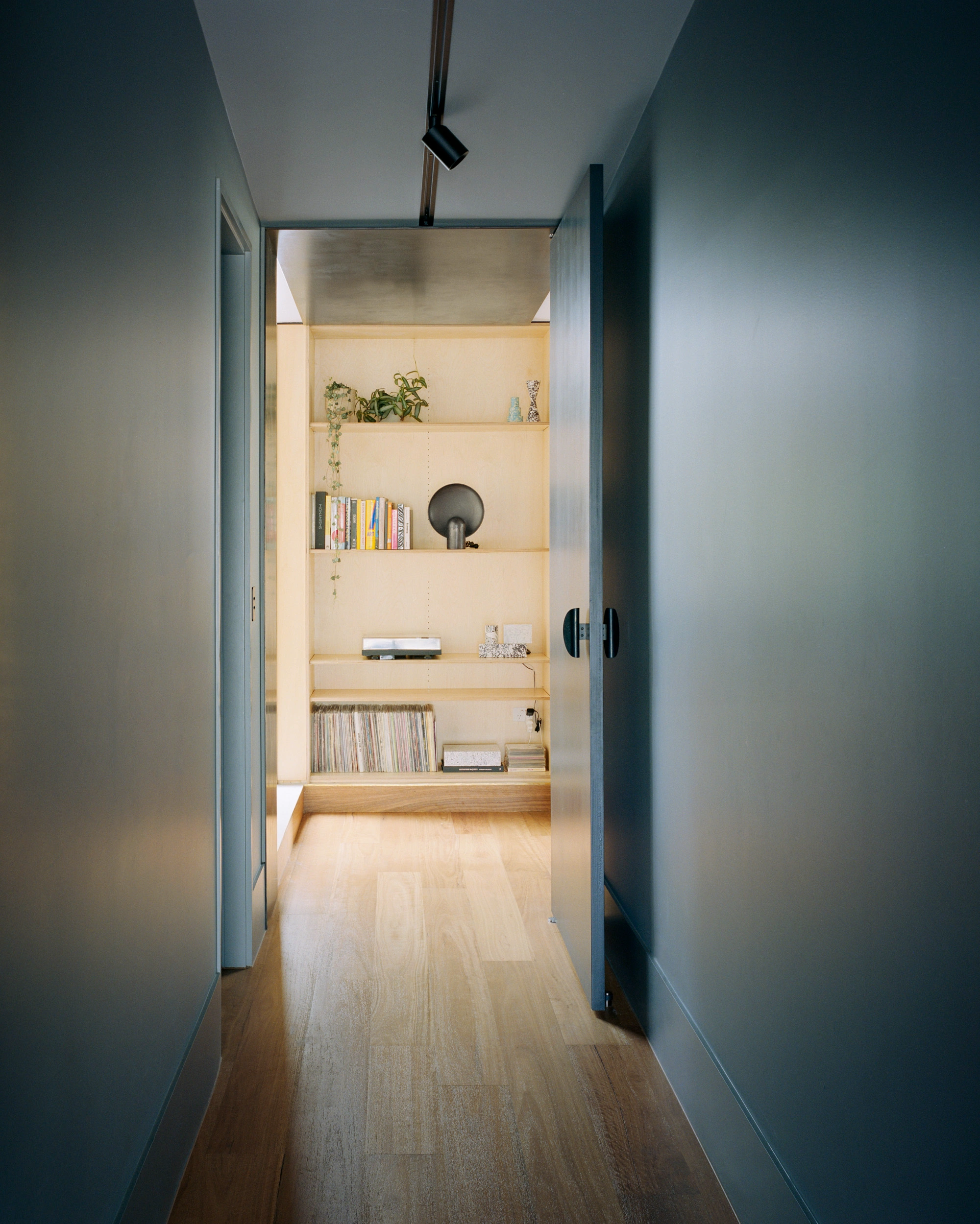 House K by Kart Projects — A view through a narrow hallway with charcoal-grey walls frames a timber-lined storage alcove beyond.