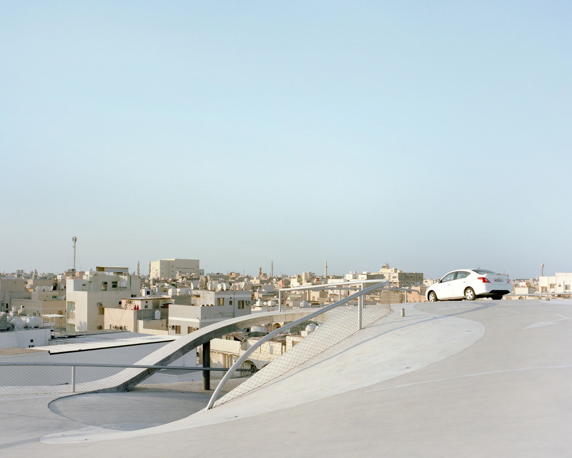 Pearling Path's Four Car Parks by Christian Kerez — A white Hyundai sedan ascends the sloping concrete ramp of Kerez's car park structure, its form silhouetted against Muharraq's dense urban skyline.