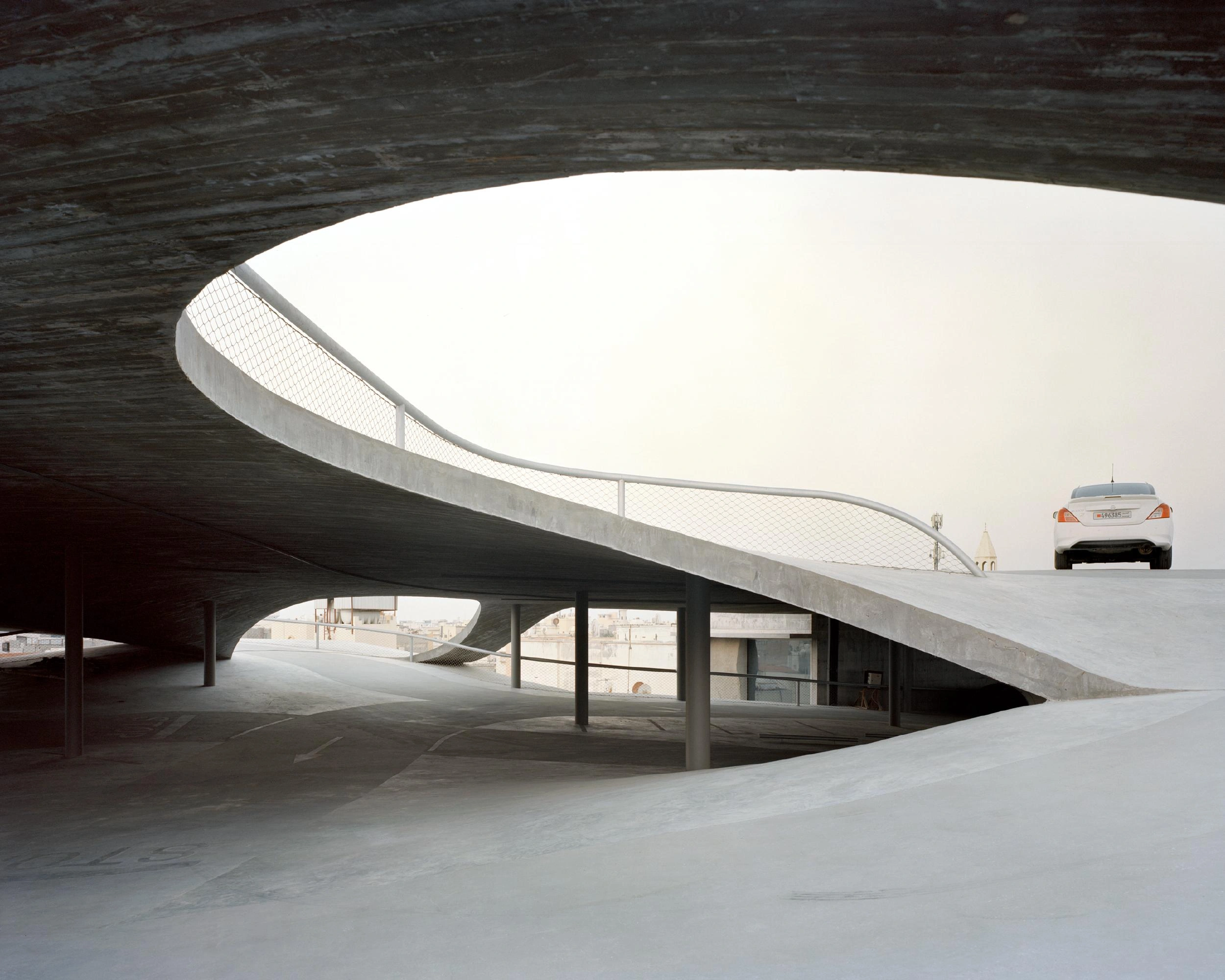 Pearling Path's Four Car Parks by Christian Kerez — A white sedan traverses the sloped concrete surface above, framed through the cavernous void of the ground-level plaza.