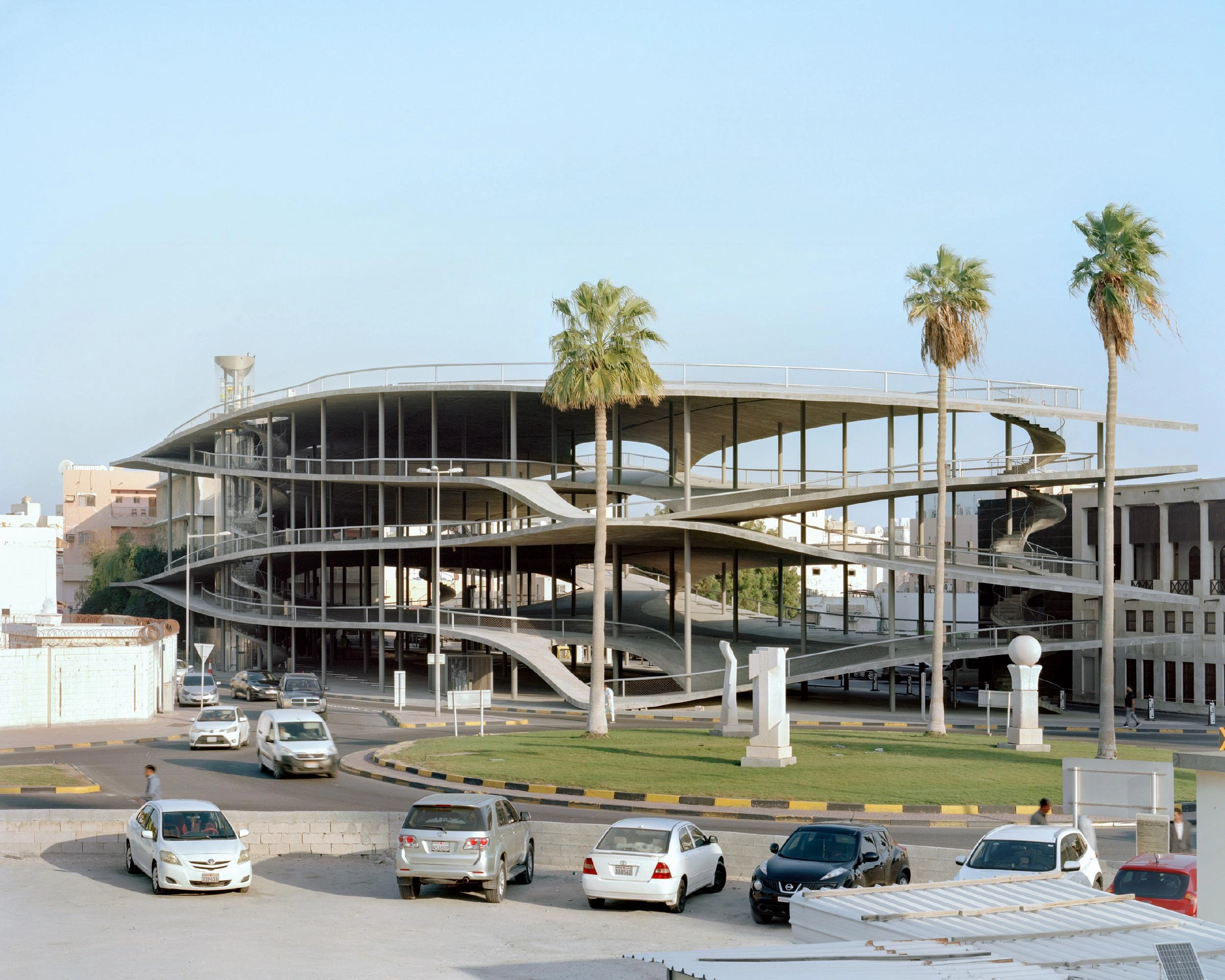Pearling Path's Four Car Parks by Christian Kerez — A spiraling concrete parking structure dominates the frame, its monolithic form punctuated by rectangular apertures that reveal interior ramp systems.