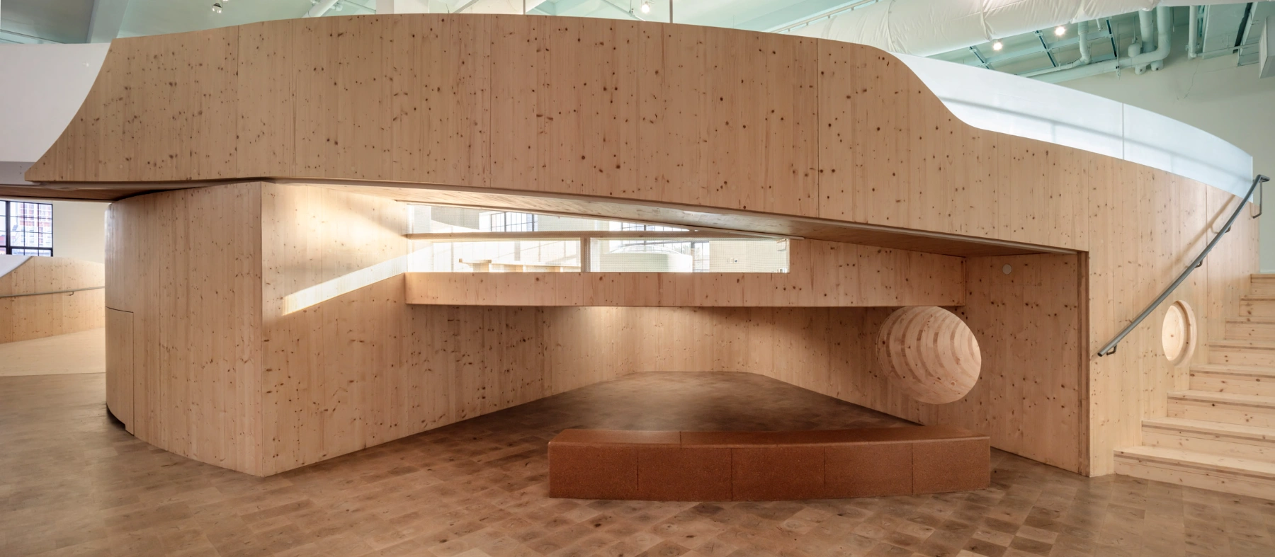Bronx Children's Museum by O'Neill McVoy — Within the timber-clad interior, a sculptural void punctures the cork-composite wall, its spiral tunnel carved to expose concentric growth rings.