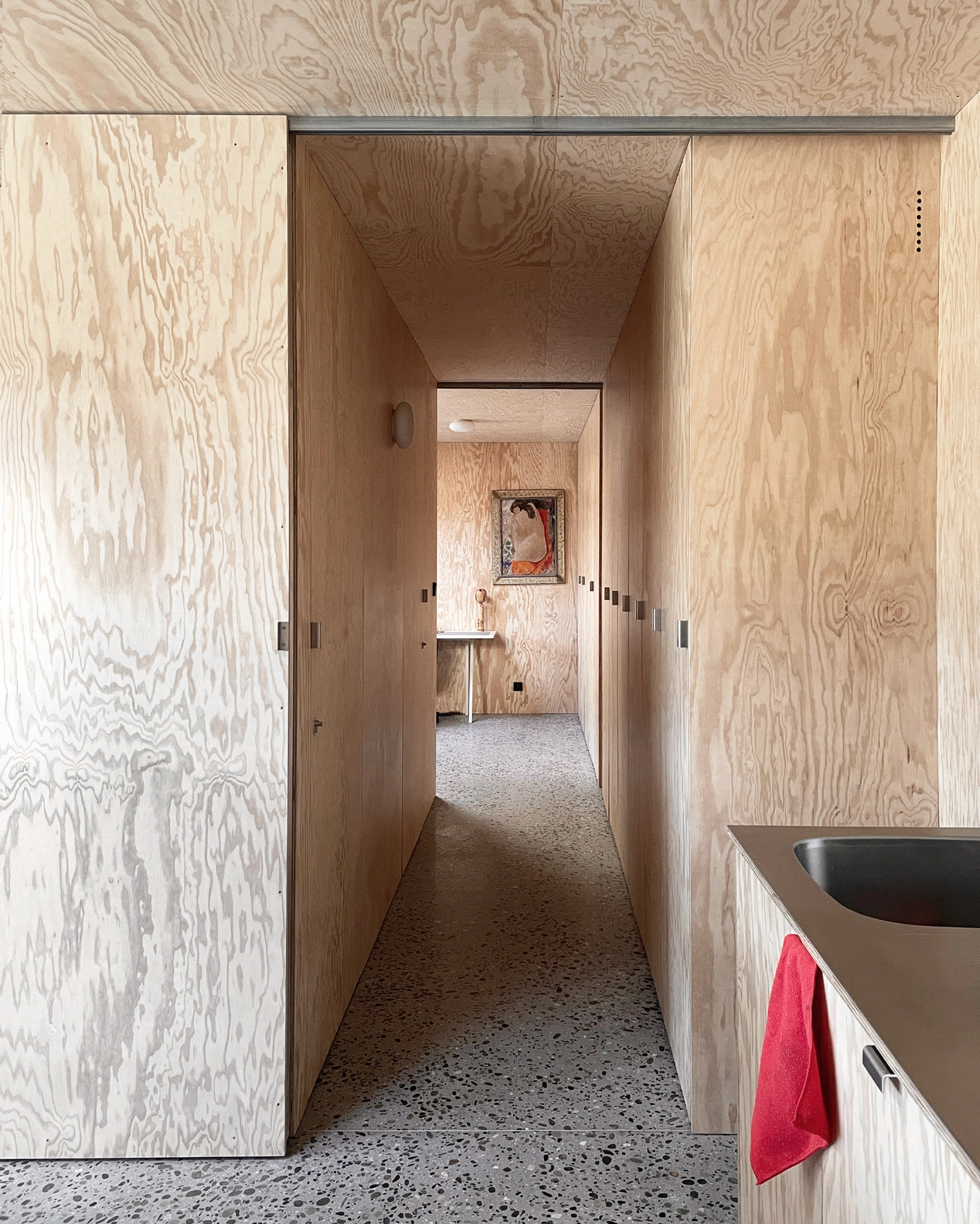 M37 House by BAST + littoral — A narrow corridor in plywood traverses the M37 House, framed by tall timber-clad walls that guide the eye toward a distant room.