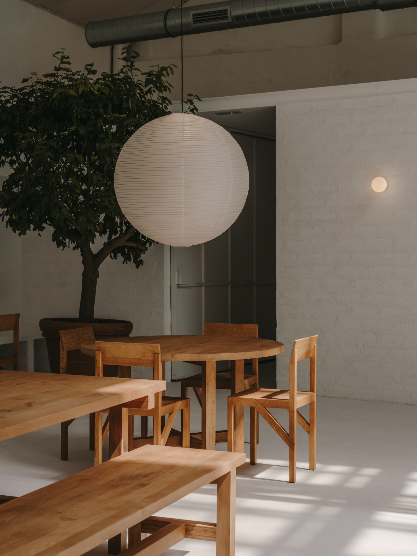 Alma Nomad by Plantea Studio by Plantea Estudio — A dining zone anchored by a round oak table with matching wooden chairs arranged in loose formation beneath a suspended paper lantern pendant.