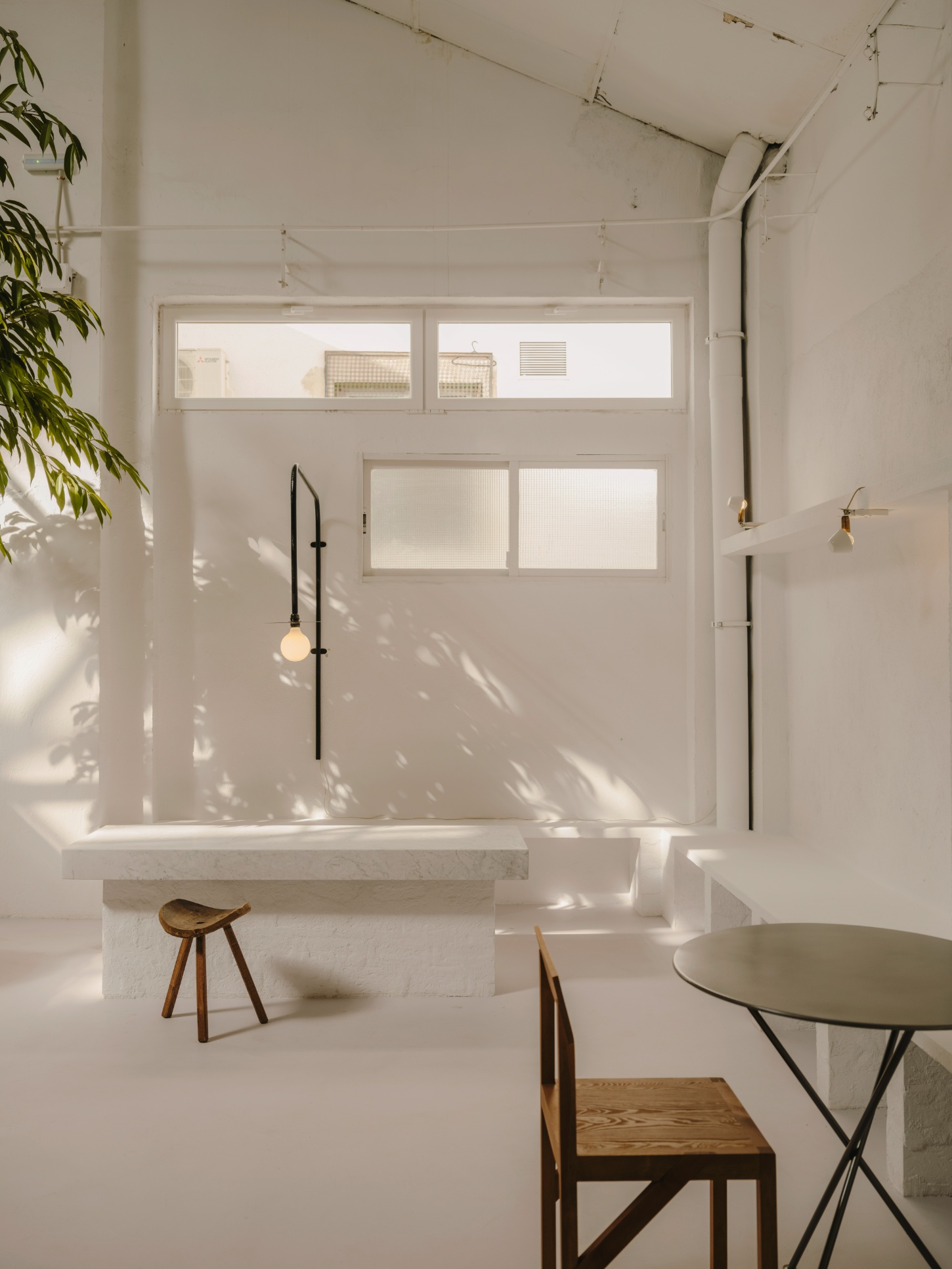 Alma Nomad by Plantea Studio by Plantea Estudio — A minimalist interior bathed in bright daylight streaming through clerestory windows, creating sharp geometric shadows across white walls and polished...