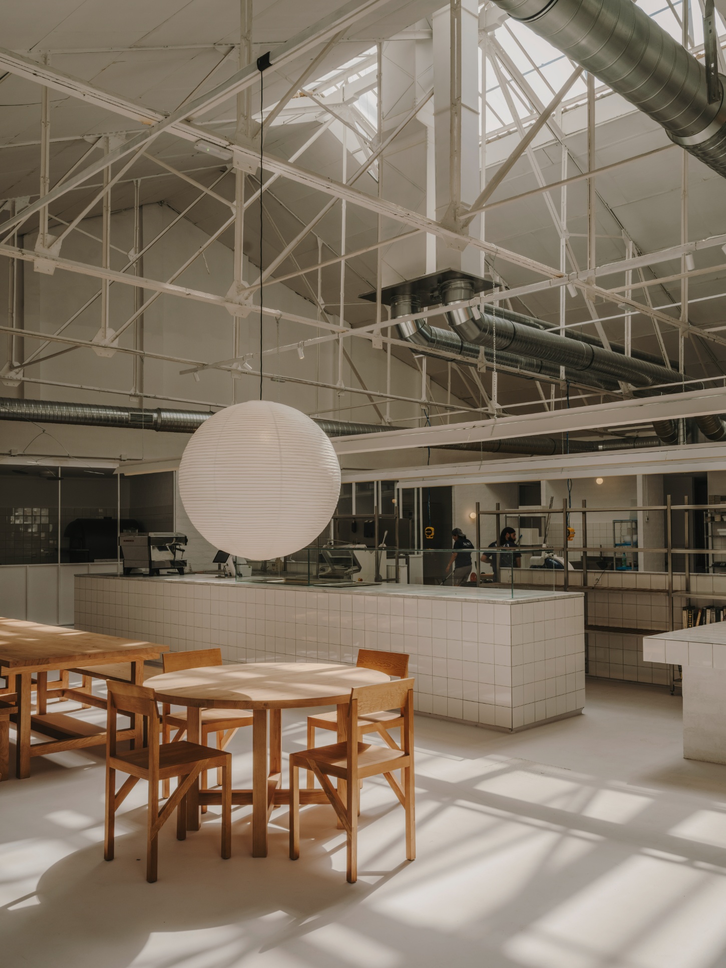 Alma Nomad by Plantea Studio by Plantea Estudio — A dining area anchored by light oak tables and chairs positioned beneath a suspended spherical pendant with horizontal ribbed detailing.