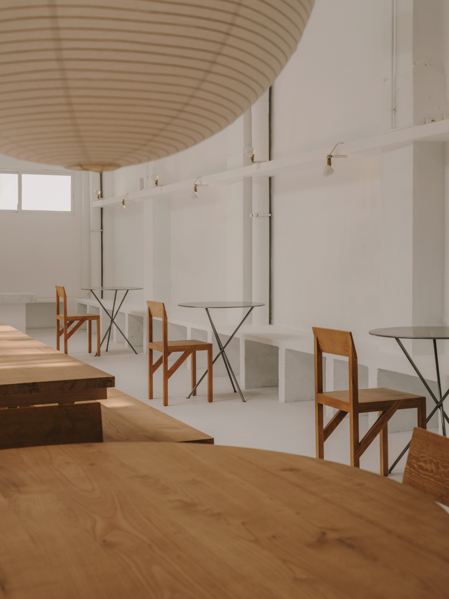 Alma Nomad by Plantea Studio by Plantea Estudio — Three round steel-framed dining tables with light oak chairs are positioned in loose clusters across a polished concrete floor.