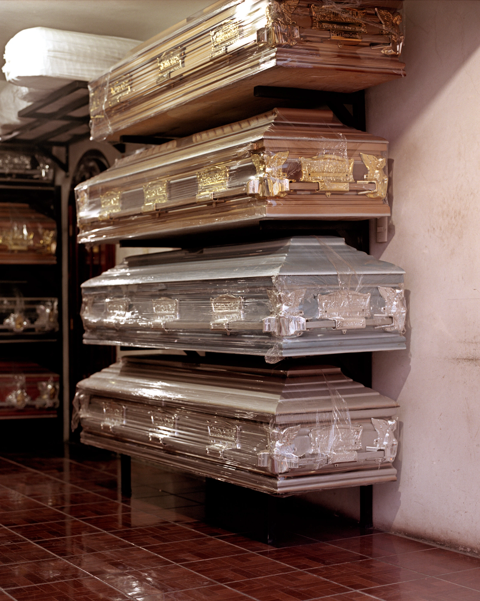 Genesis by Juan Brenner — Stacked wooden caskets with brass hardware displayed on industrial metal shelving against a mauve-toned plaster wall.