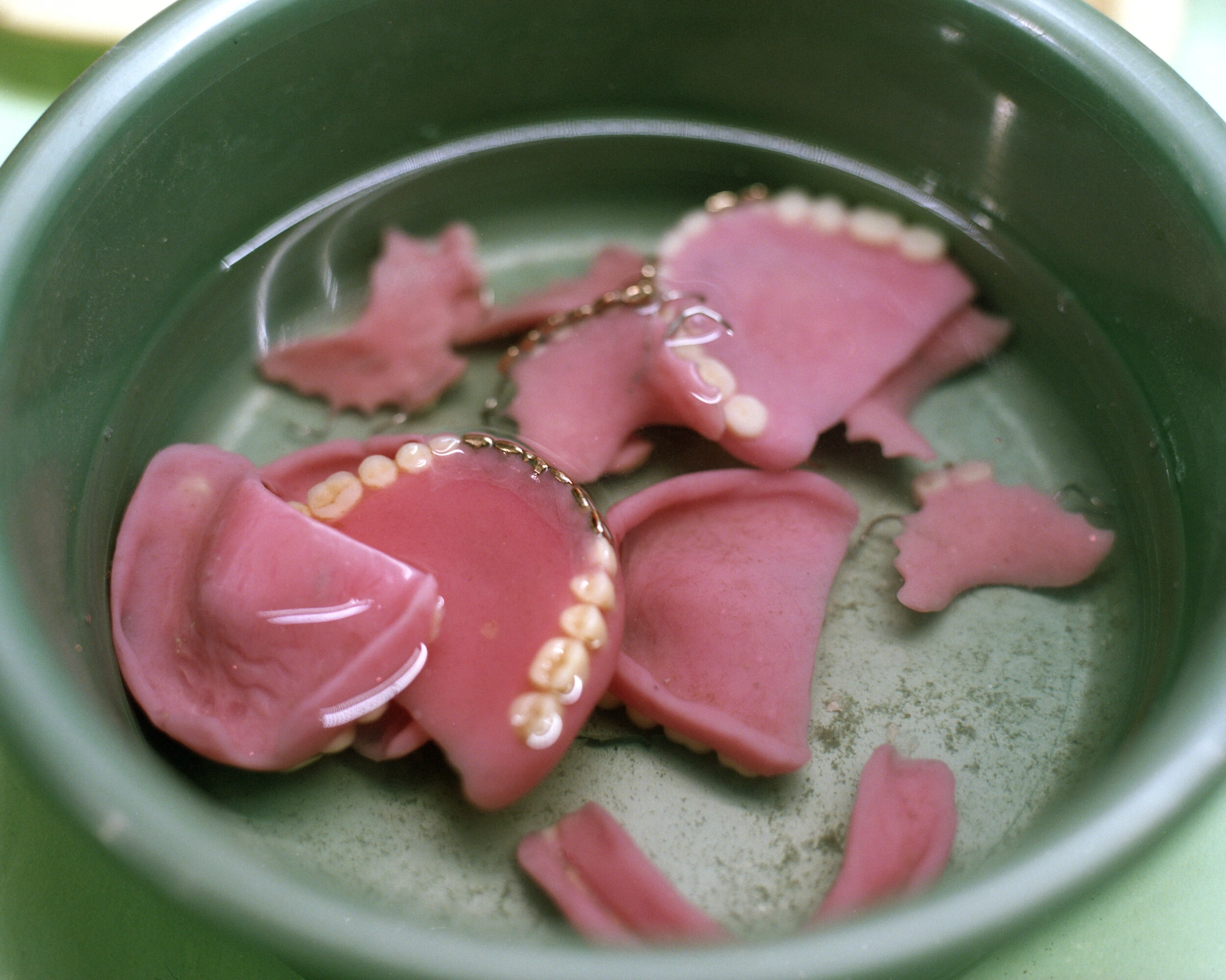 Genesis by Juan Brenner — Pink resin charms submerged in shallow water within a ceramic vessel, their surfaces detailed with white pearl beads and gold chain accents.