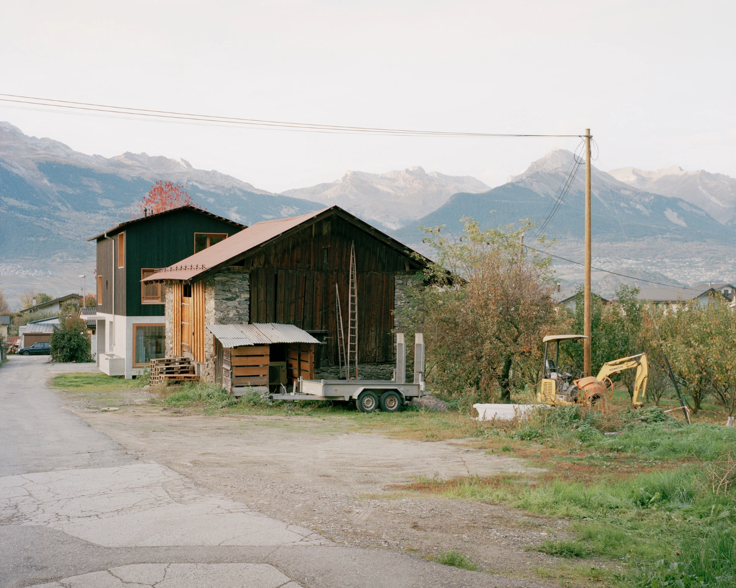 The Wandering House by Lionel Ballmer — A contemporary timber-clad volume elevated on a white concrete plinth sits adjacent to a dark-stained wooden barn structure.
