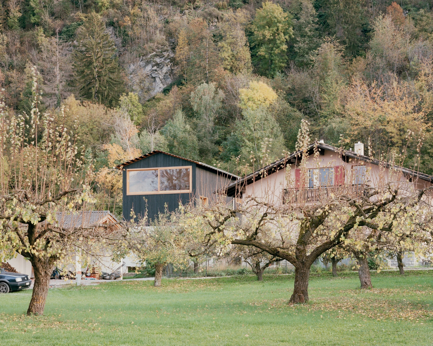 The Wandering House by Lionel Ballmer — A pitched-roof structure with charred timber vertical cladding dominates the composition, its dark facade punctuated by a continuous horizontal window band...