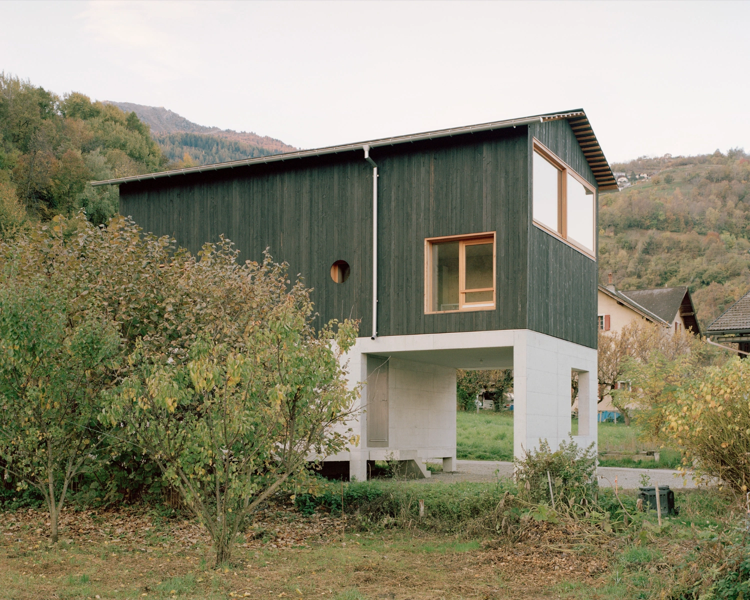 The Wandering House by Lionel Ballmer — A two-story residential volume clad in charred vertical timber boards rises above a white concrete plinth, creating a stark material contrast.