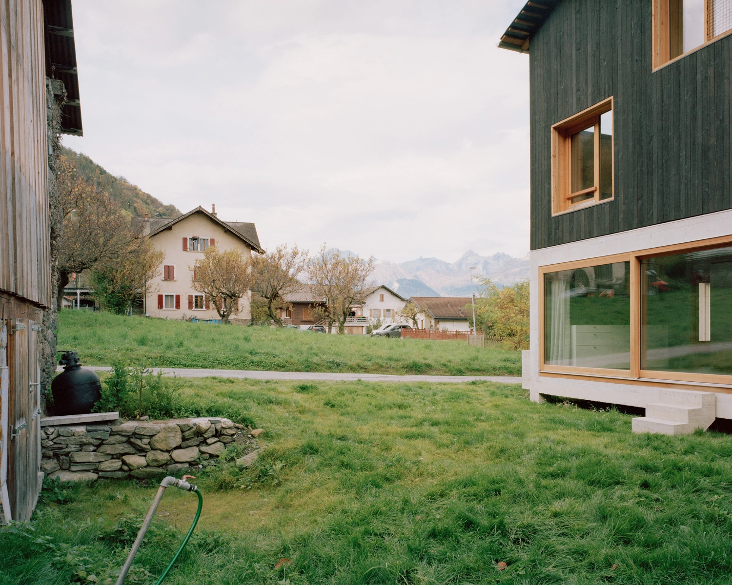 The Wandering House by Lionel Ballmer — The green-clad timber volume dominates the right edge of frame, its corrugated siding interrupted by a continuous horizontal window band with timber surround.