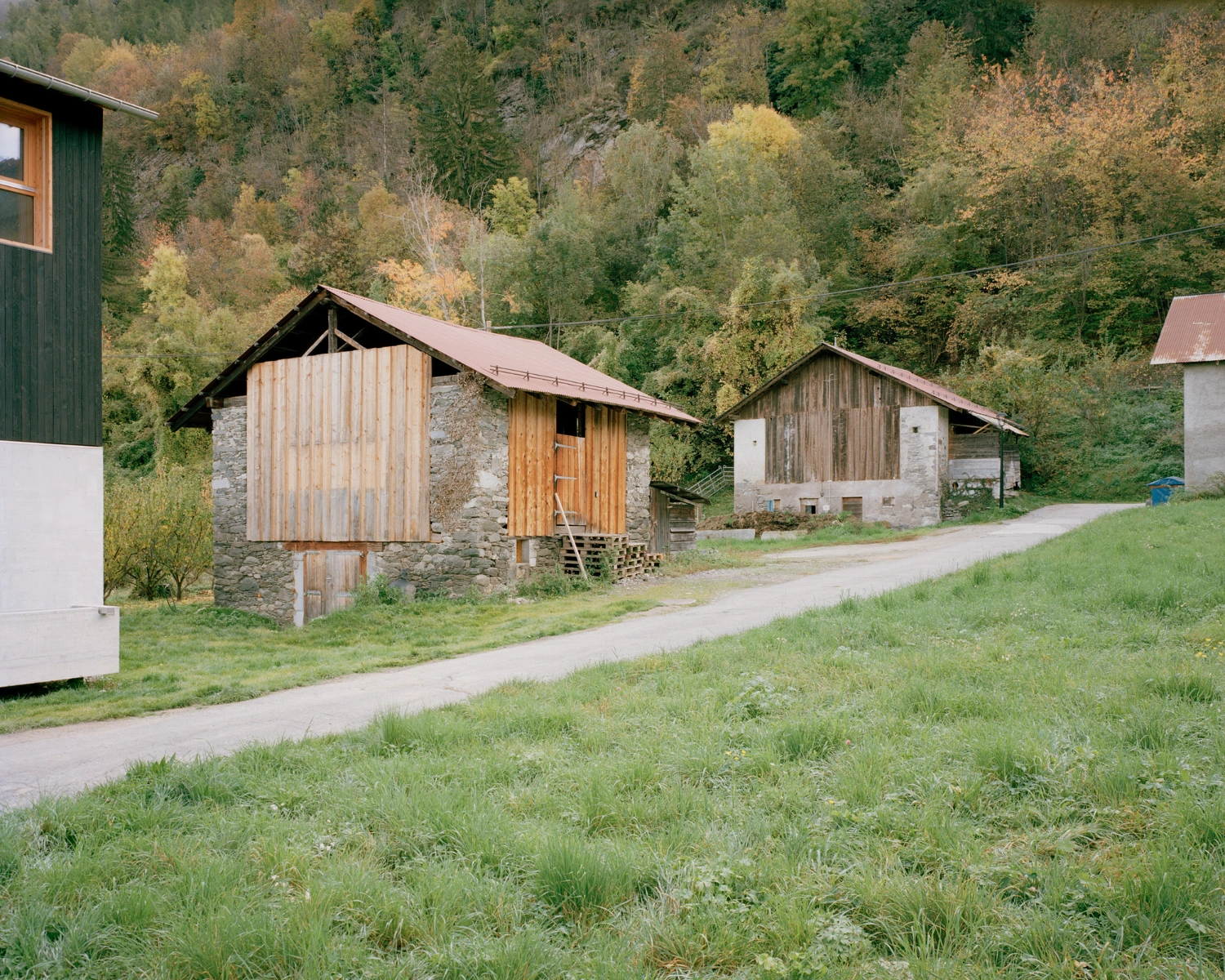 The Wandering House by Lionel Ballmer — Two timber-clad structures occupy a sloped alpine clearing, their weathered wood siding arranged in vertical boards showing distinct patination patterns.
