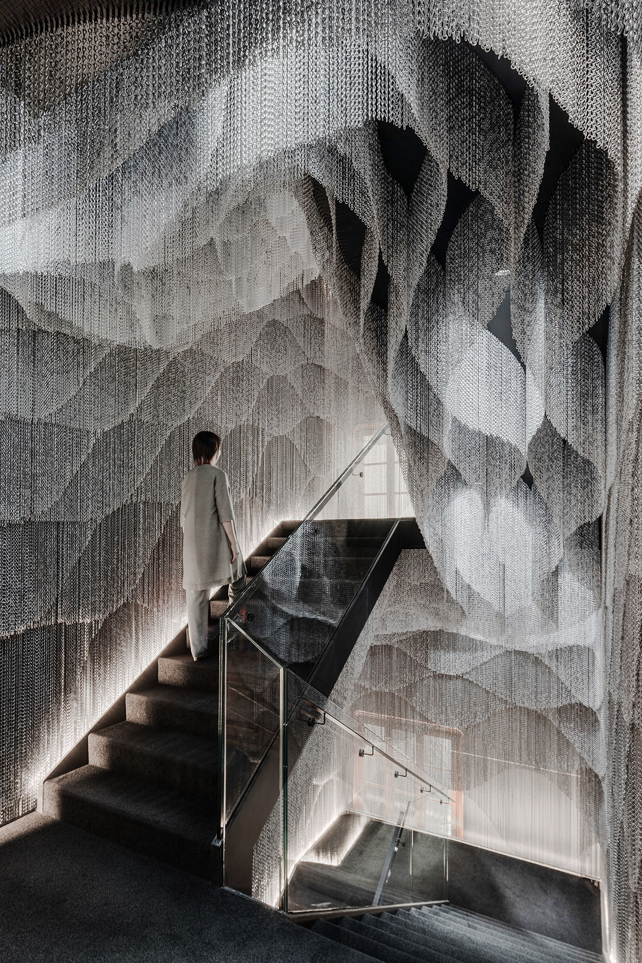 Casa Batlló Stairs & Atrium by Kengo Kuma — A solitary figure ascends a concrete staircase enclosed by floor-to-ceiling cascading aluminum chain curtains that fragment natural light into a spectrum of...