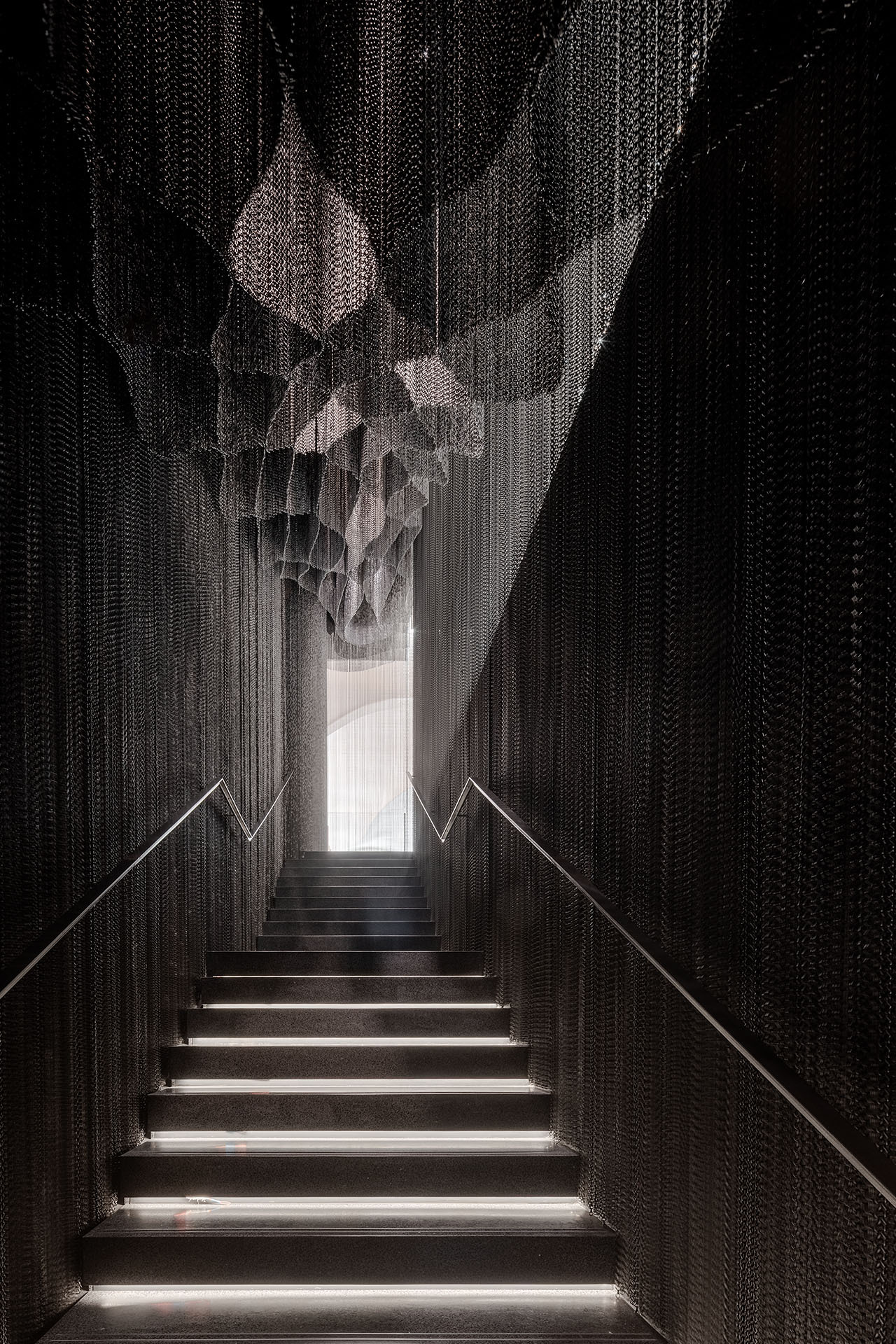 Casa Batlló Stairs & Atrium by Kengo Kuma — A narrow concrete staircase descends into shadow, flanked by cascading aluminum chain curtains that envelop both walls from floor to ceiling.