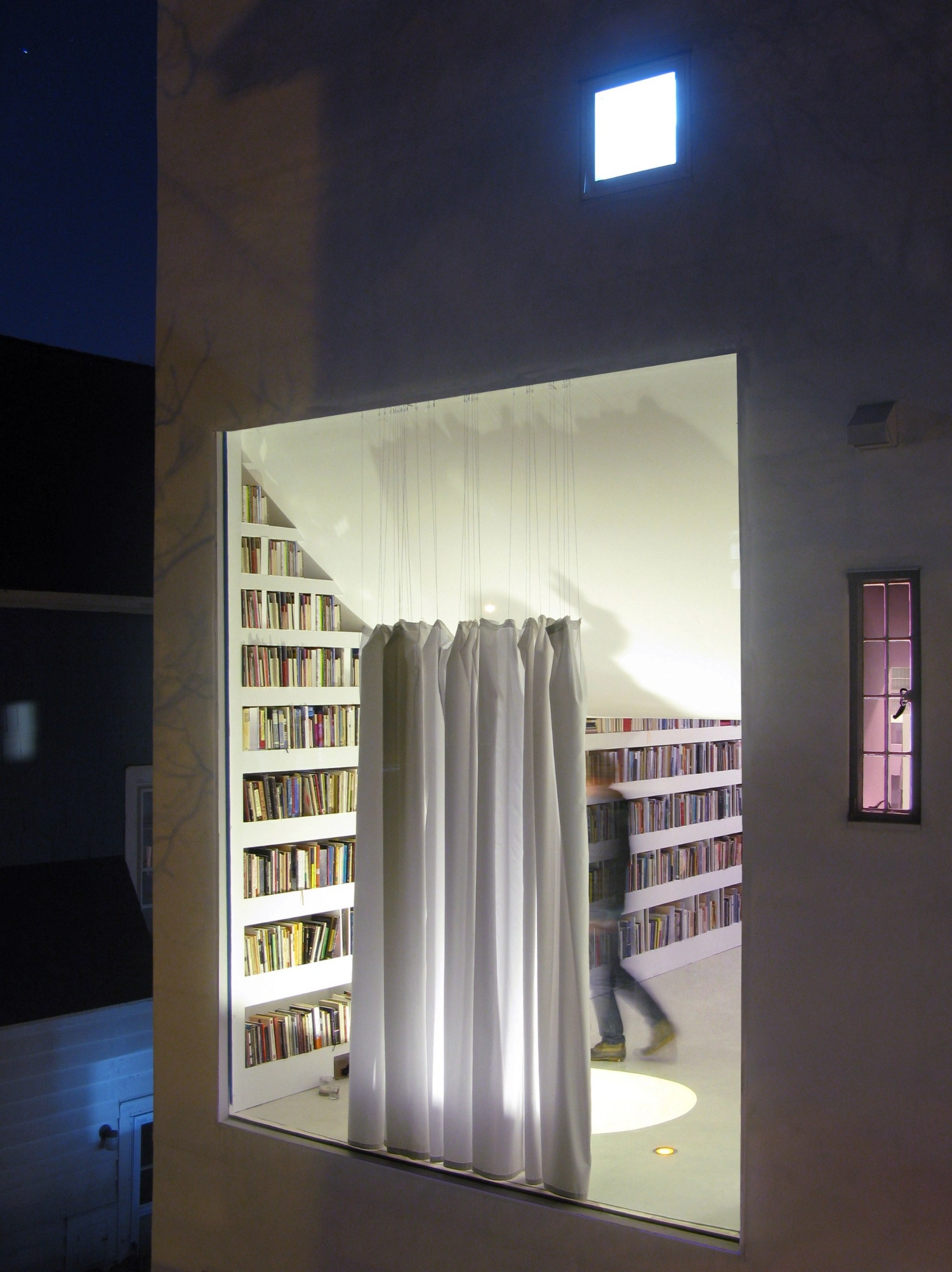 Haffenden House by PARA — A glowing rectangular aperture interrupts the dark facade of Haffenden House, revealing stacked bookshelves and a billowing curtain system that diffuses...