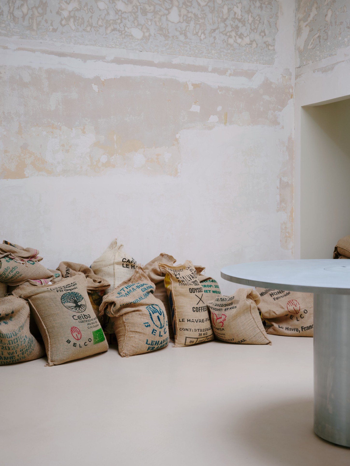 Anbassa by Office Abrami Rojas by Office Abrami Rojas (OAR) — Burlap coffee sacks stamped with origin text and certification marks—Le Havre, France; Belco; various weights and dates—are arranged on a white table surface.