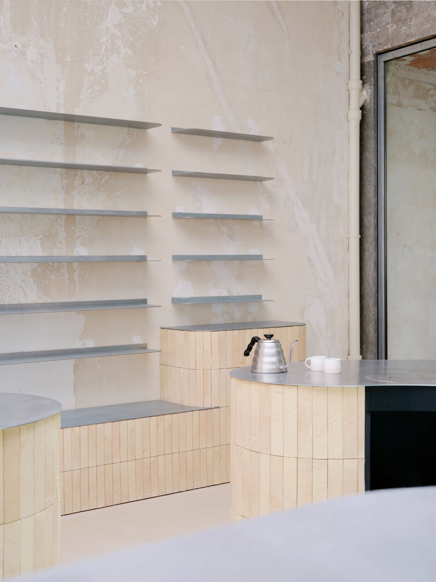 Anbassa by Office Abrami Rojas by Office Abrami Rojas (OAR) — A coffee service sits atop a curved timber counter clad in vertical pine slats, metal-framed shelving mounted above displaying industrial steel brackets.