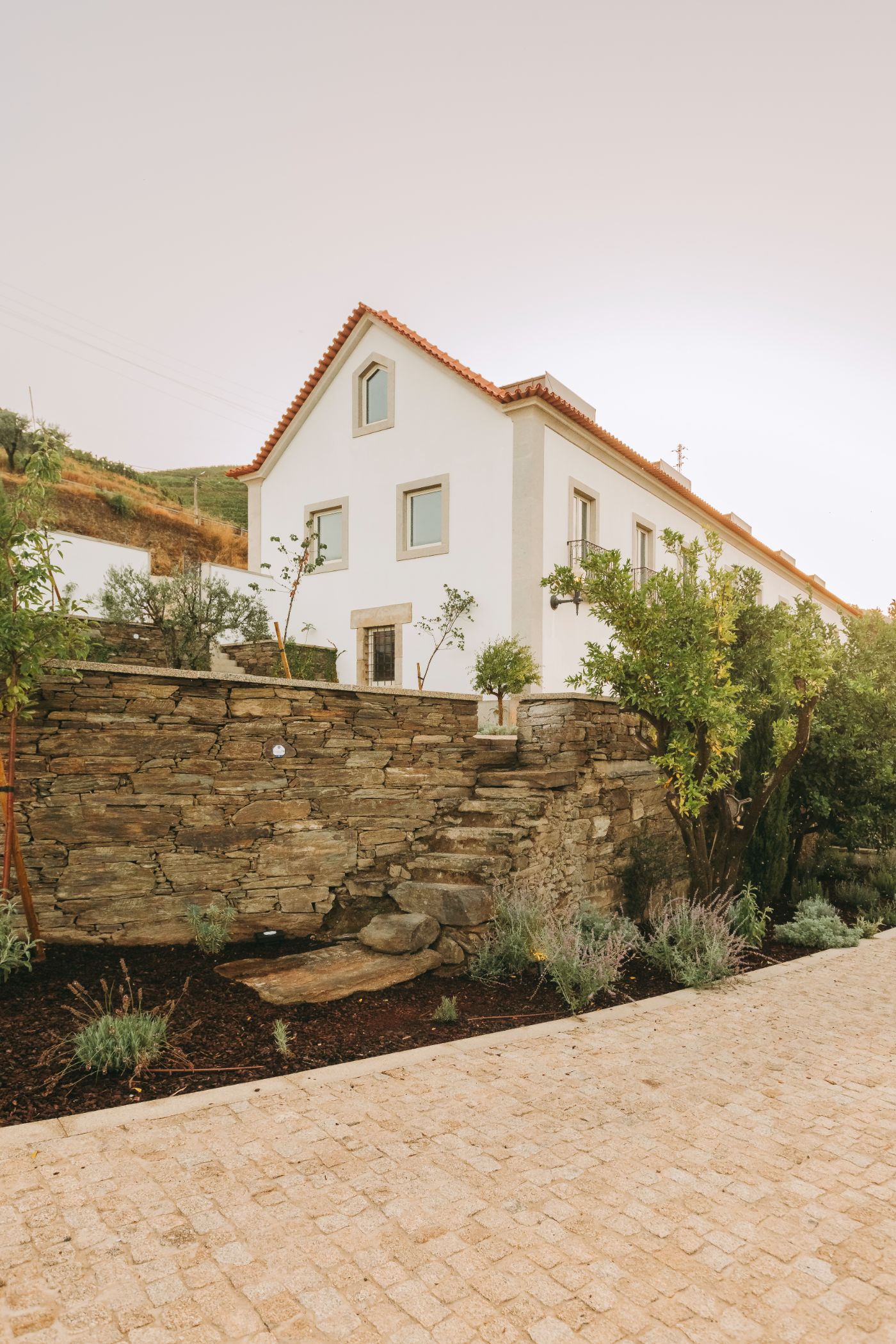 Torel Quinta da Vacaria in Douro Valley by Studio Astolfi and architect Luís Miguel Oliveira — A rendered cream-colored villa with terracotta clay tile roof sits elevated behind a dry-stacked stone retaining wall.