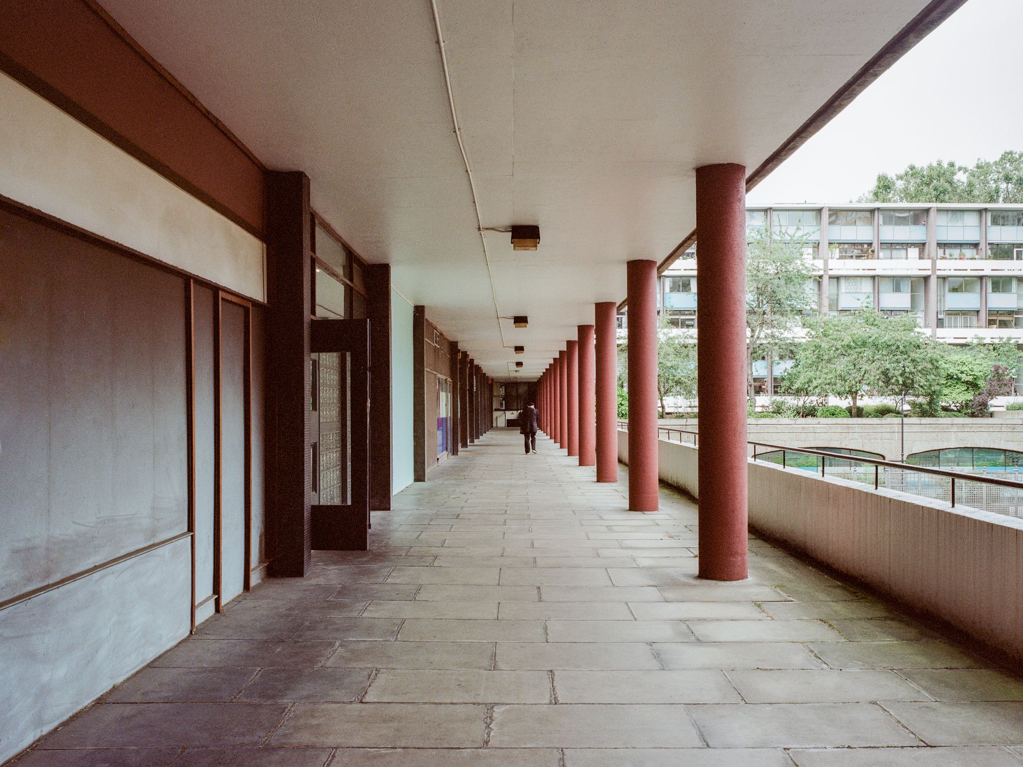Crescent House by TYPE — A covered passage with regular structural rhythm defined by cylindrical columns in deep burgundy, receding toward distant figures.