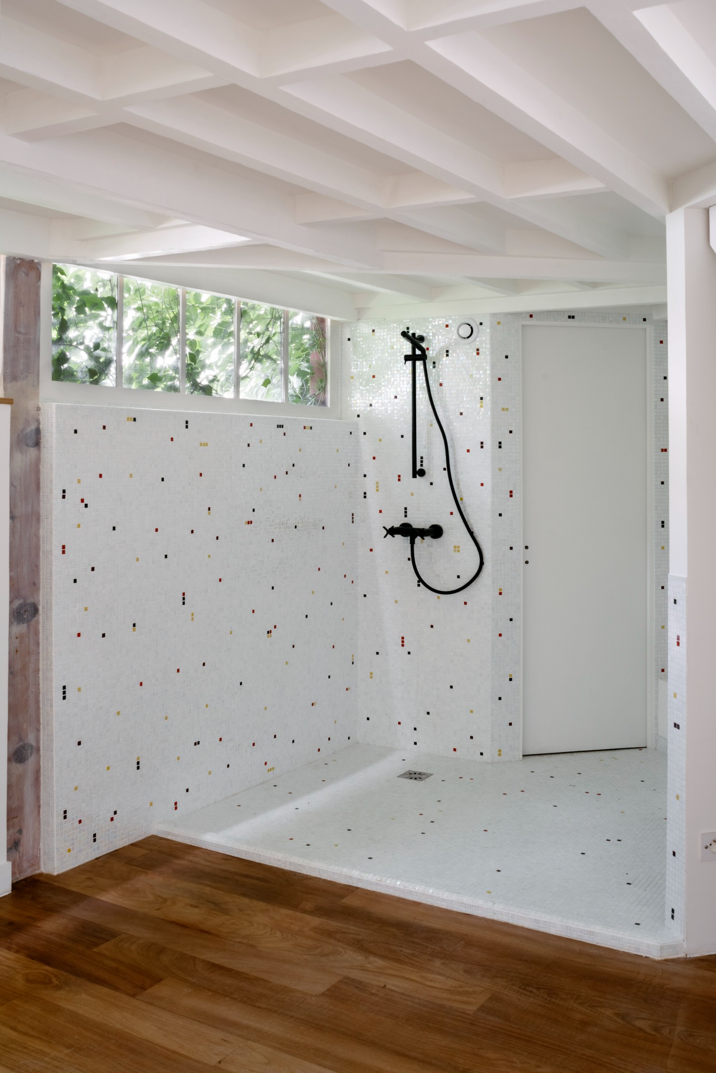 Atelier 22 by Atelier Mula Scala + Atelier Mas by Atelier Mura Scala and Atelier Mas — A walk-in shower enclosure lined with white ceramic tile punctuated by scattered small tiles in black, red, and gold.