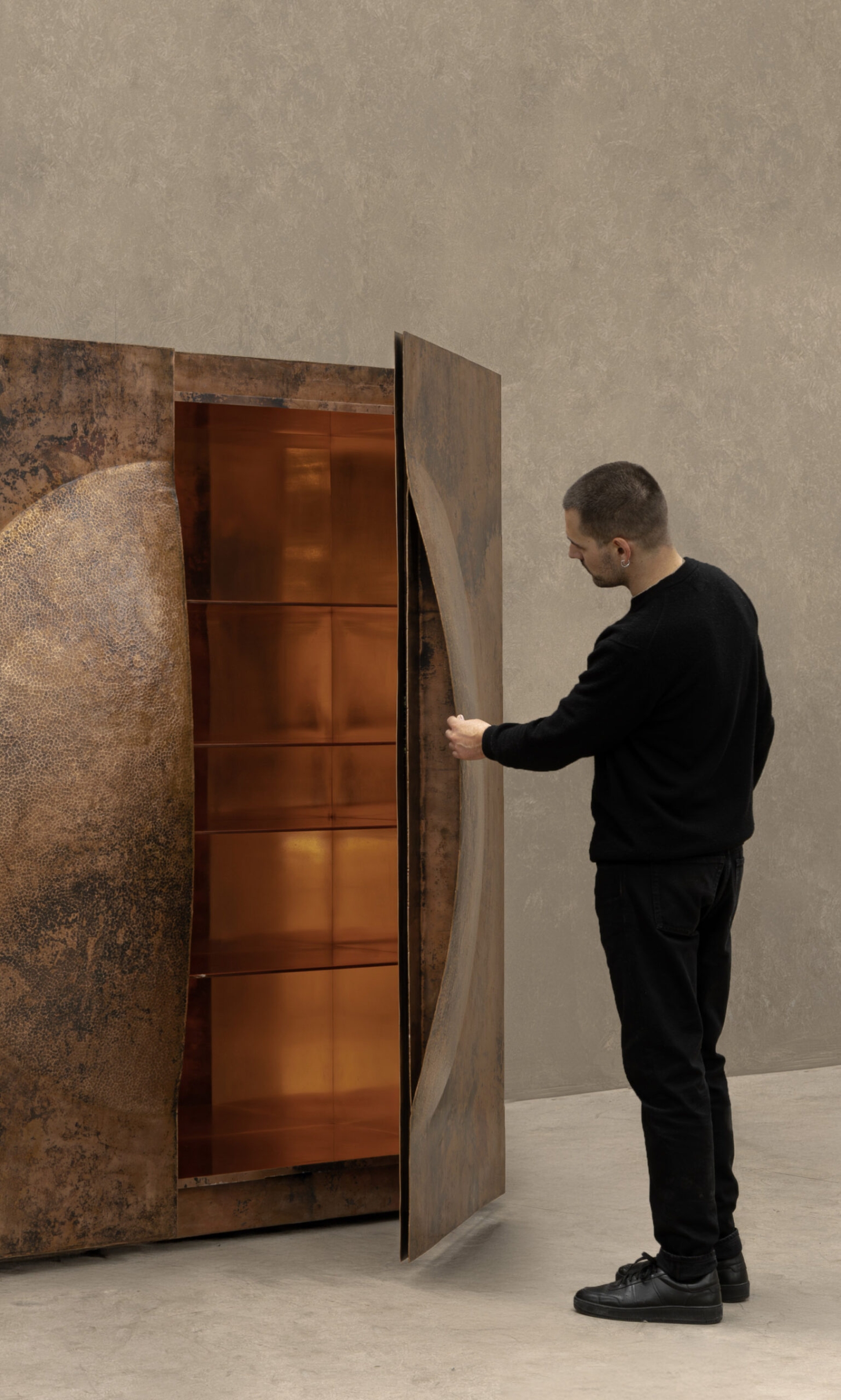 New Works by Manu Bañó — A man in black clothing stands before a copper cabinet, its left face displaying a textured, oxidized patina in deep rust-brown tones.