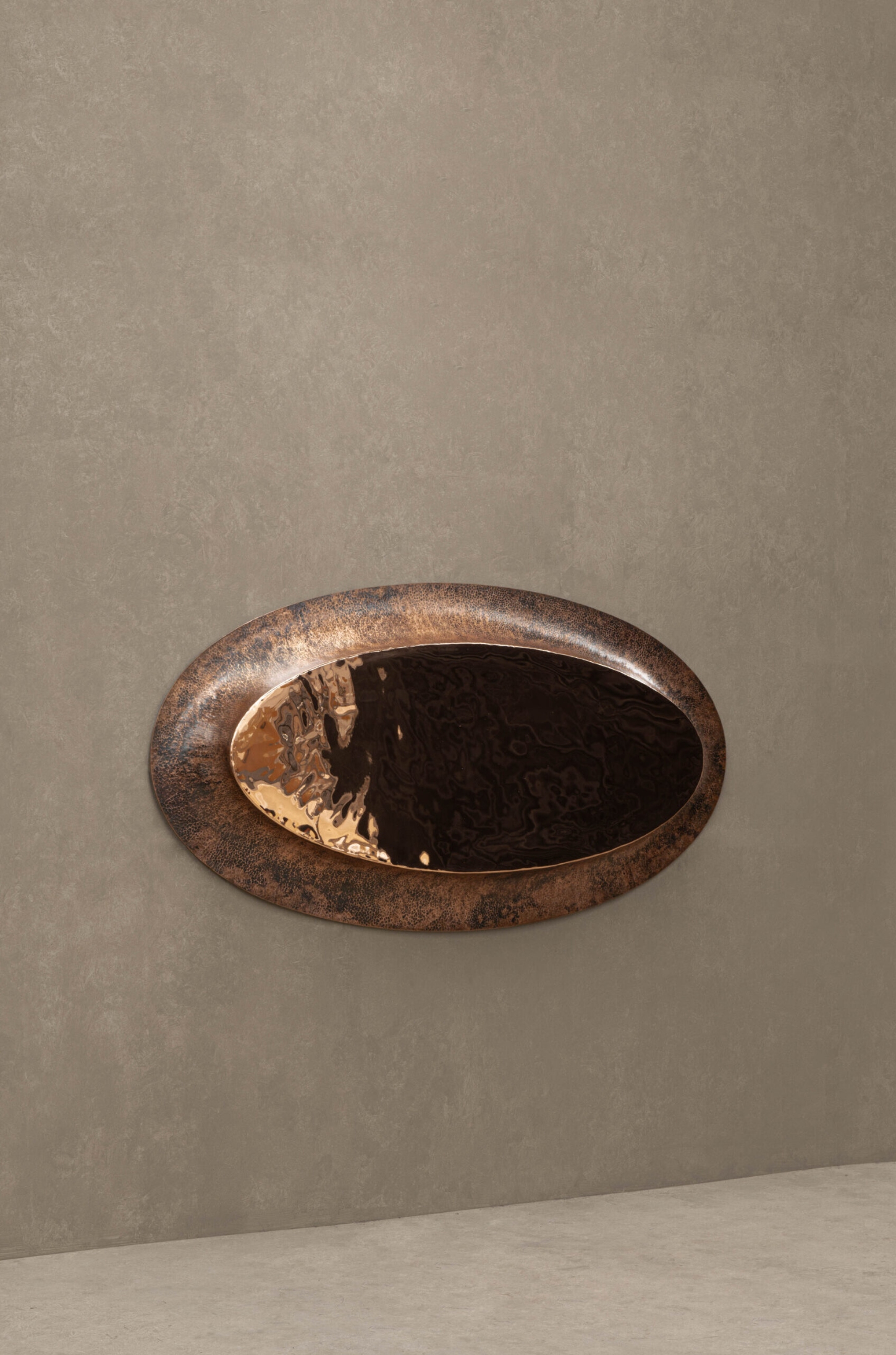 New Works by Manu Bañó — A horizontal oval mirror with oxidized copper frame mounted on a neutral wall.