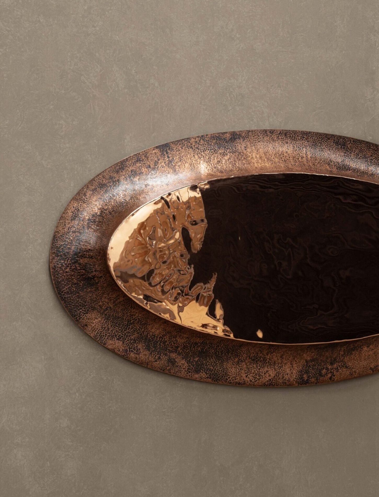 New Works by Manu Bañó — An oval mirror with oxidized copper frame dominates the composition, its surface divided by a vertical seam into two symmetrical halves.
