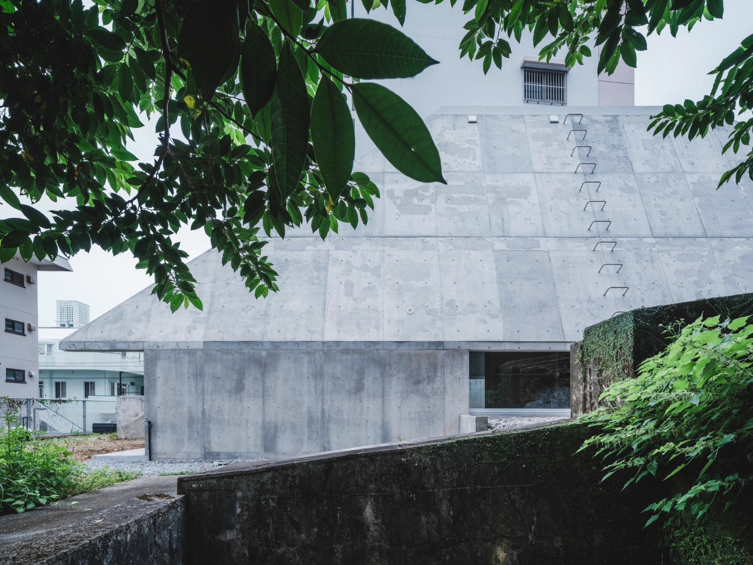 Pyramid Hut by IGArchitects — Framed through overhanging foliage, the concrete volume of Pyramid Hut presents its trapezoidal facade with board-formed seams running horizontally across...