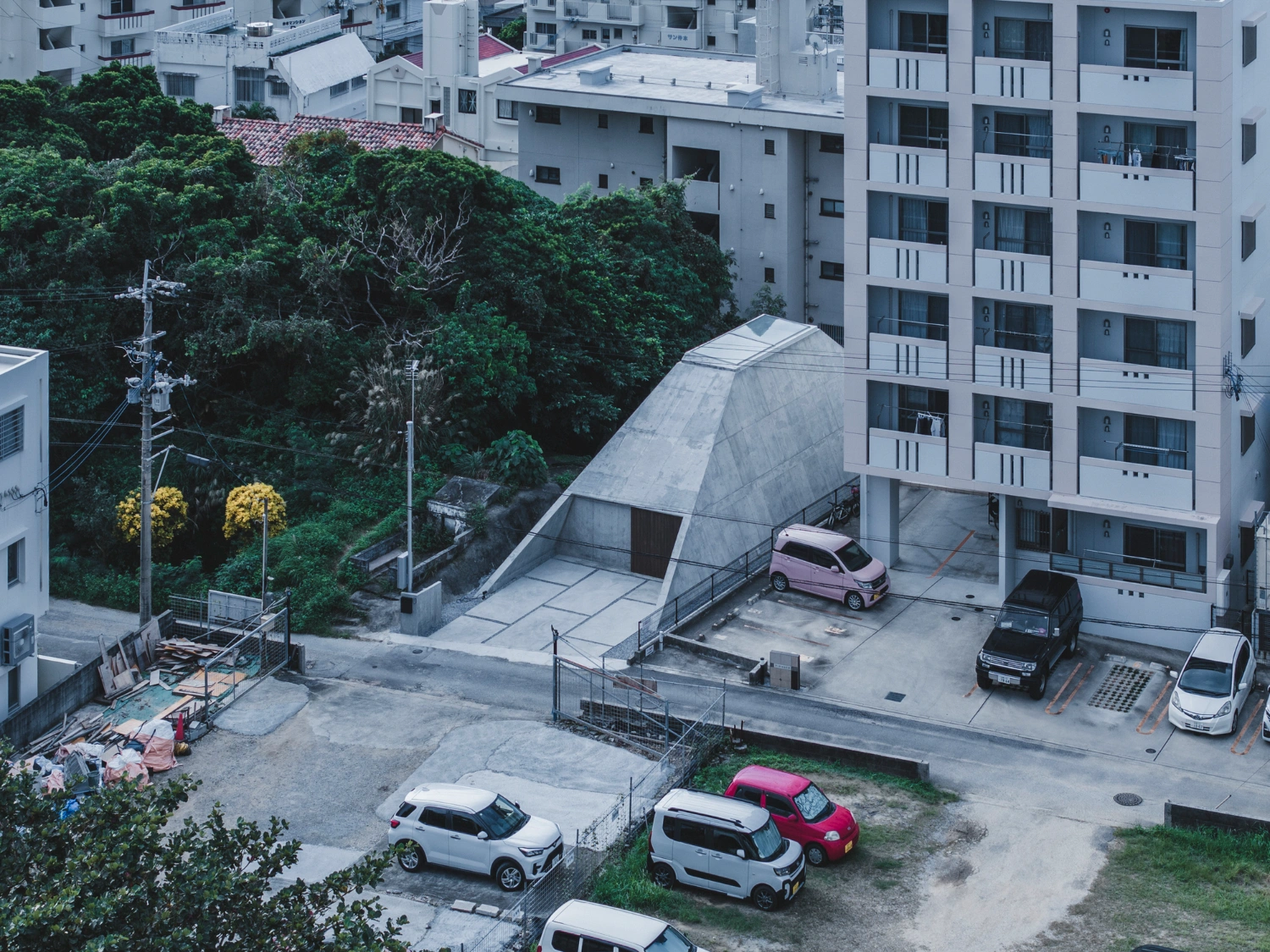 Pyramid Hut by IGArchitects — Aerial view of Pyramid Hut's forecourt and approach, revealing the trapezoidal concrete volume nestled within a dense residential context.