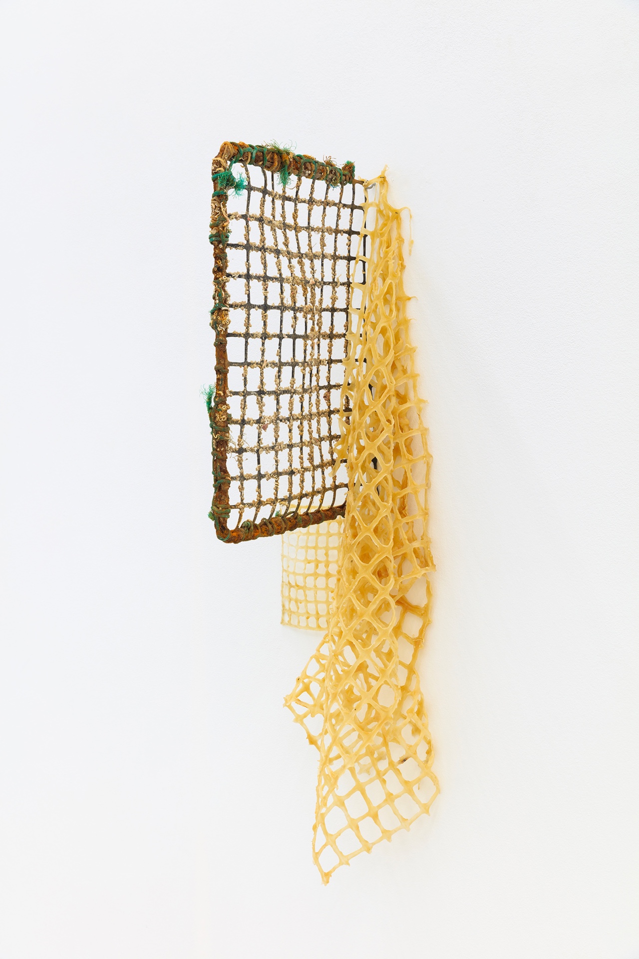 Não Faz Mal by Manon Harnois at Galeria Foco — A suspended textile installation composed of two distinct mesh panels in graduated yellows, with the upper rectangular section featuring a tightly woven...