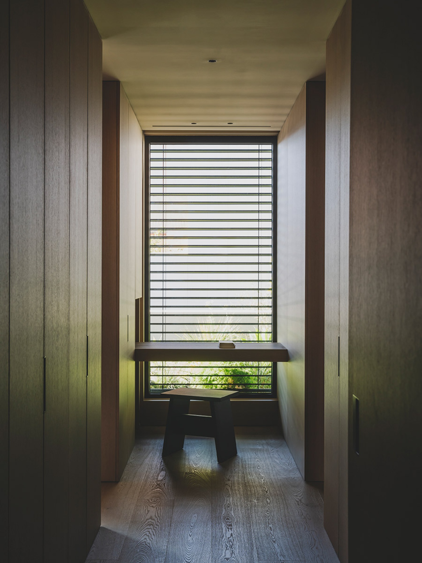 S29 House by Francesc Rifé Studio + Exit Projectes — A timber-lined corridor extends toward a large window fitted with horizontal louvered blinds, filtering daylight into horizontal striations across...