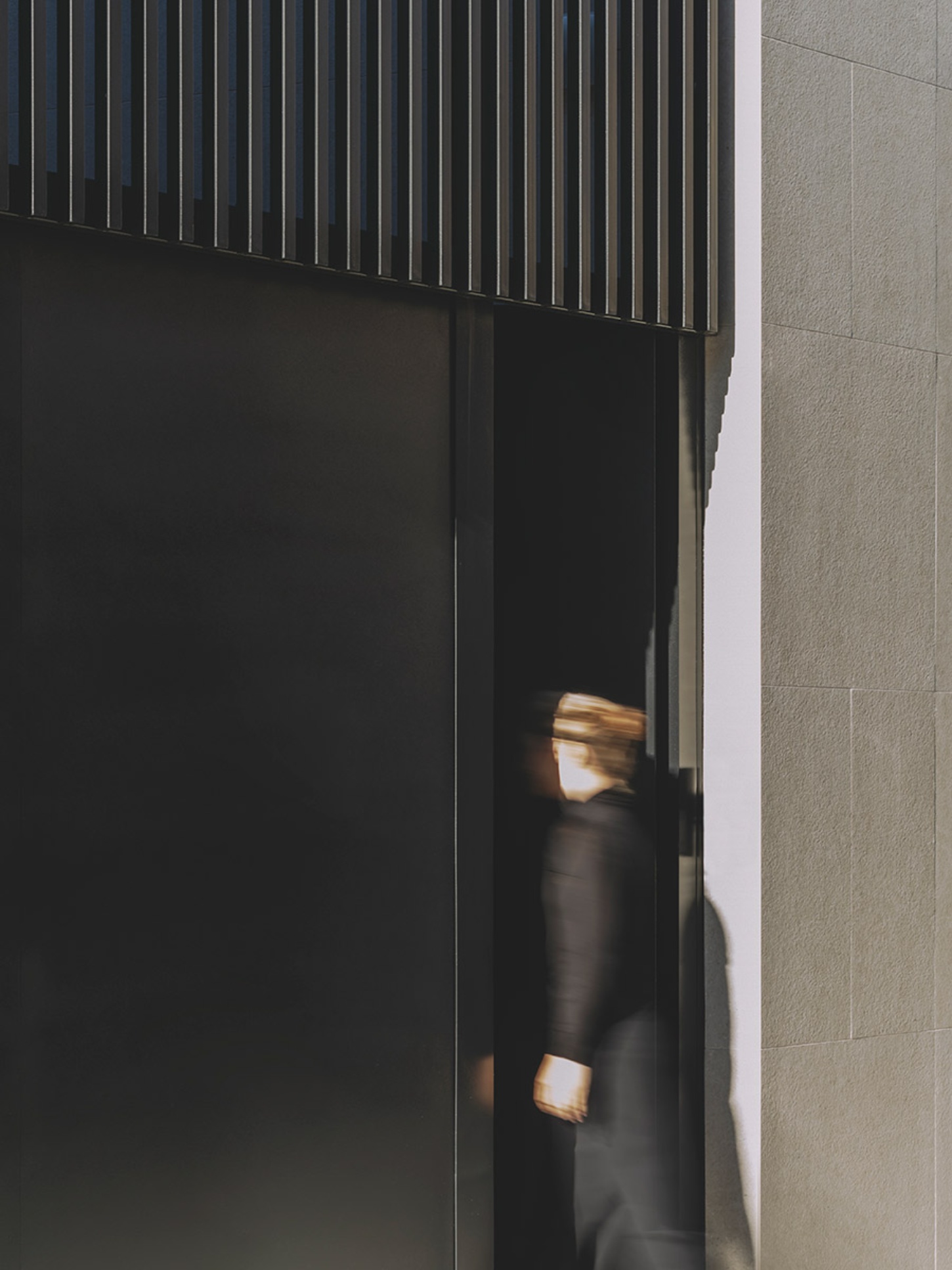 S29 House by Francesc Rifé Studio + Exit Projectes — A figure moves through a narrow passage between a dark corrugated metal panel wall and a textured stone or render surface.