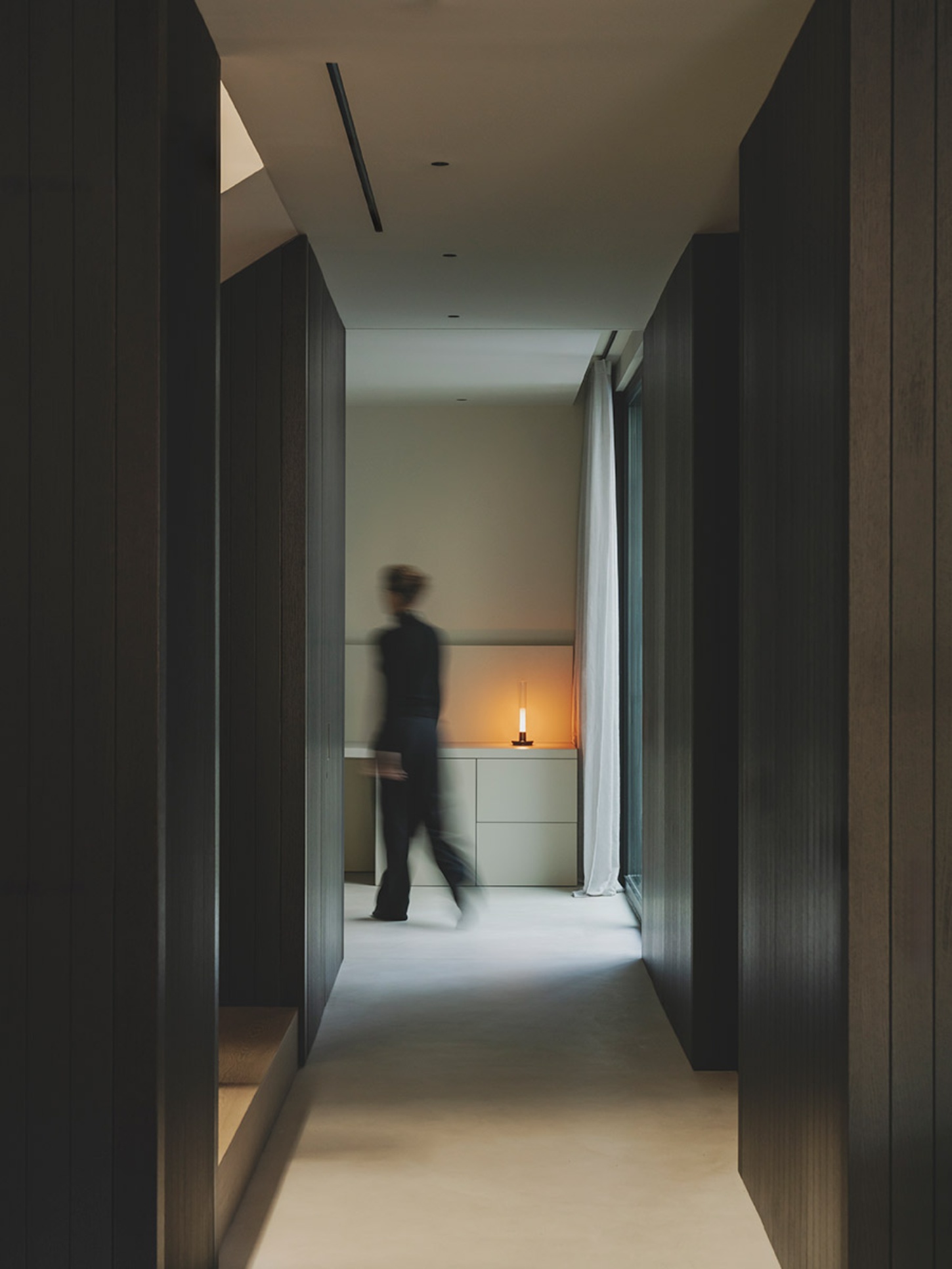 S29 House by Francesc Rifé Studio + Exit Projectes — A narrow corridor lined with dark-stained timber paneling creates a compressed spatial passage toward a luminous threshold.