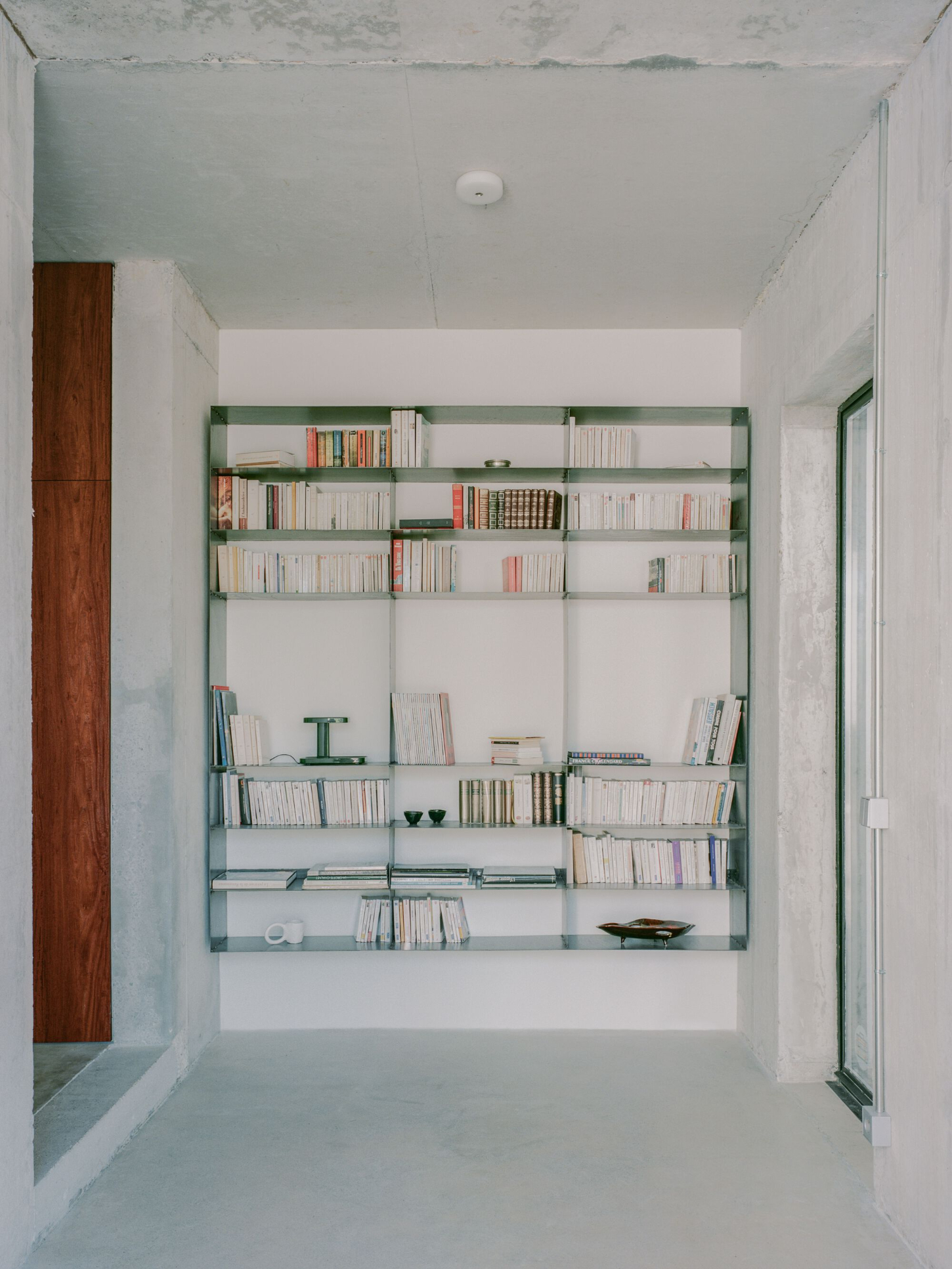 PAYZ House by Alors Studio — A built-in shelving system occupies the full height of a white-walled interior room, its frame constructed from green-painted metal with frosted glass shelving.