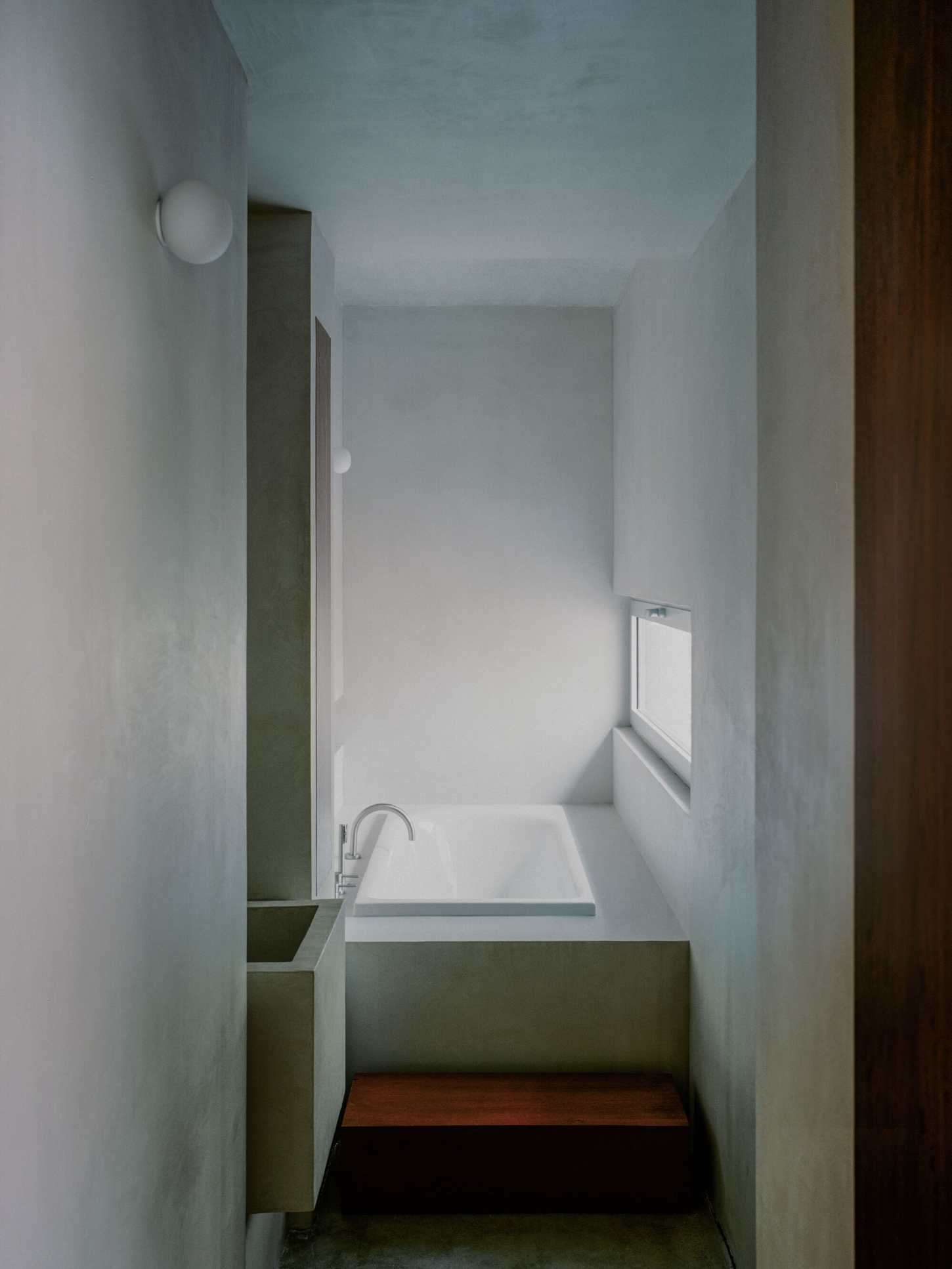 PAYZ House by Alors Studio — A narrow bathroom viewed from the threshold reveals a recessed bathtub with white surround positioned against the far wall.