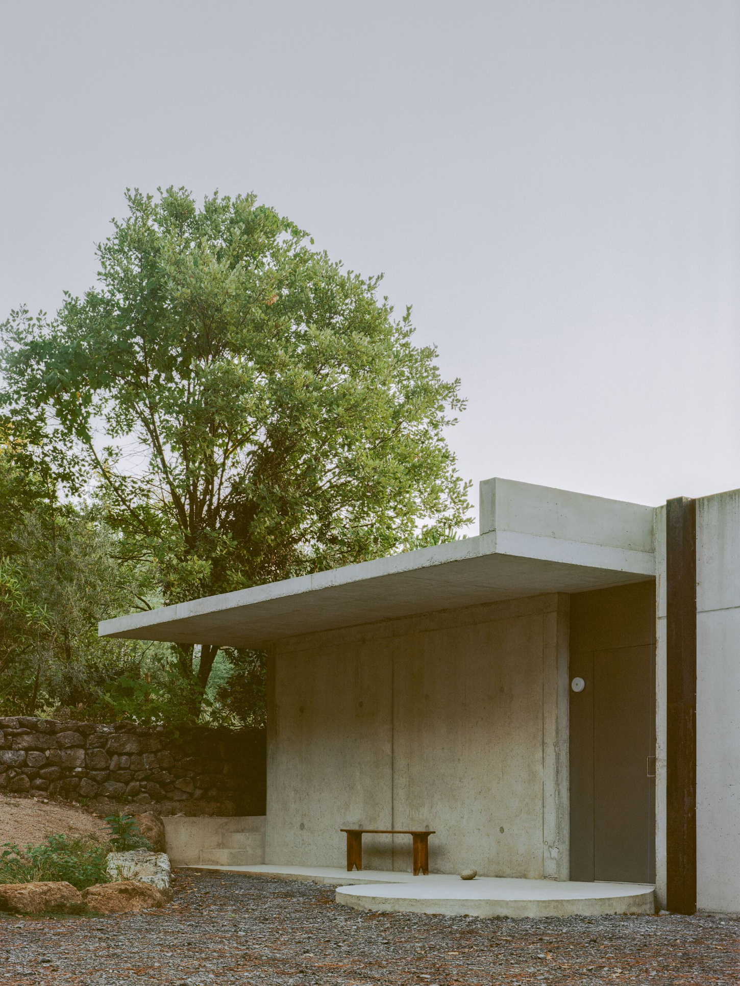 PAYZ House by Alors Studio — A monolithic concrete pavilion anchors the gravel courtyard, its board-formed facade weathered to a silvered patina.