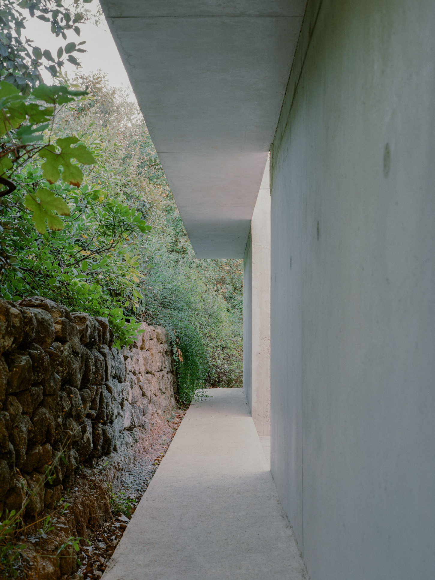 PAYZ House by Alors Studio — A narrow passage carved between a weathered dry stone wall and a monolithic concrete structure demonstrates the project's contextual insertion into the...