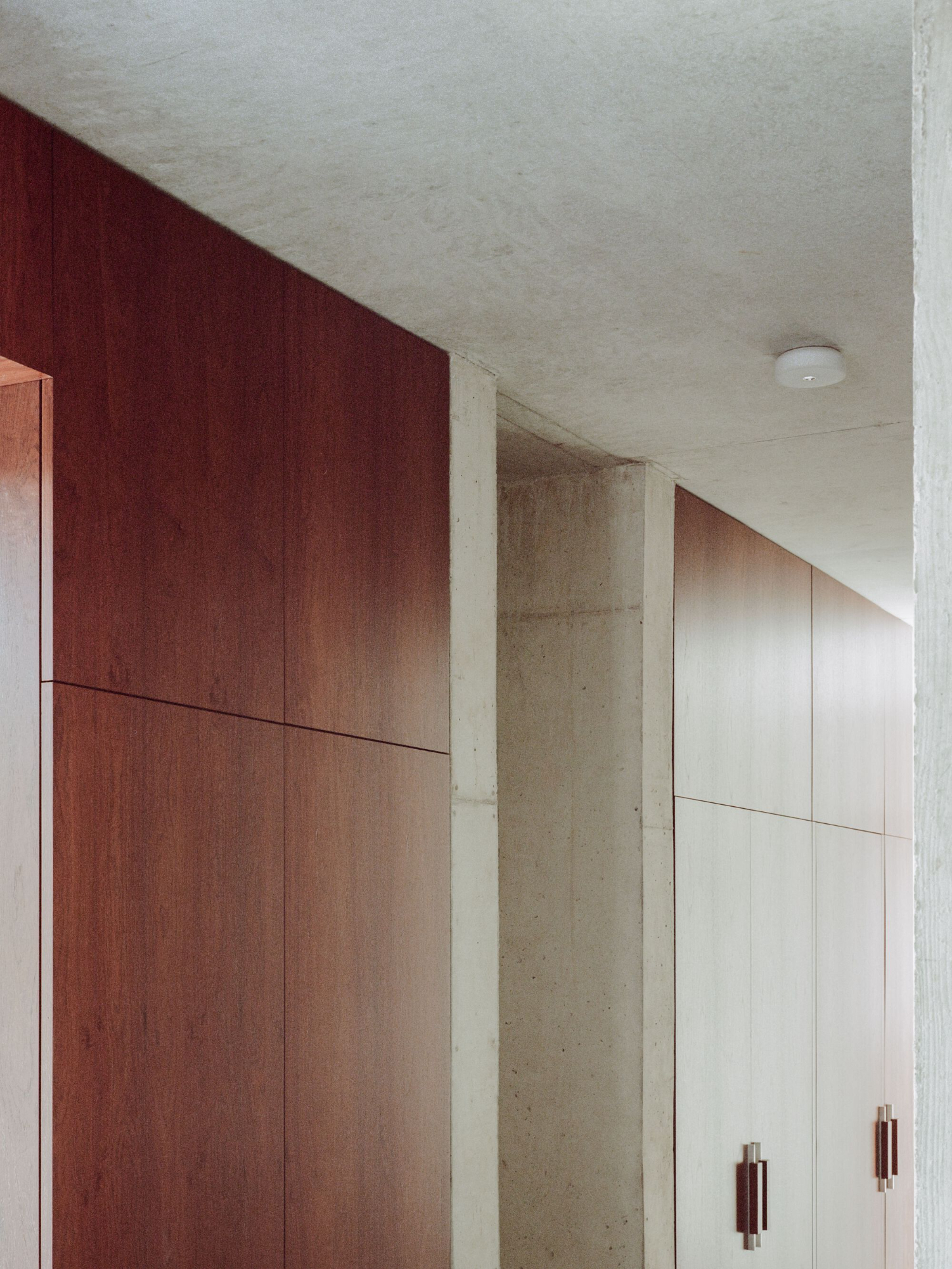 PAYZ House by Alors Studio — A corridor lined with built-in cabinetry in contrasting finishes—deep burgundy wood paneling on the left, cream-colored panels on the right—separated by a...