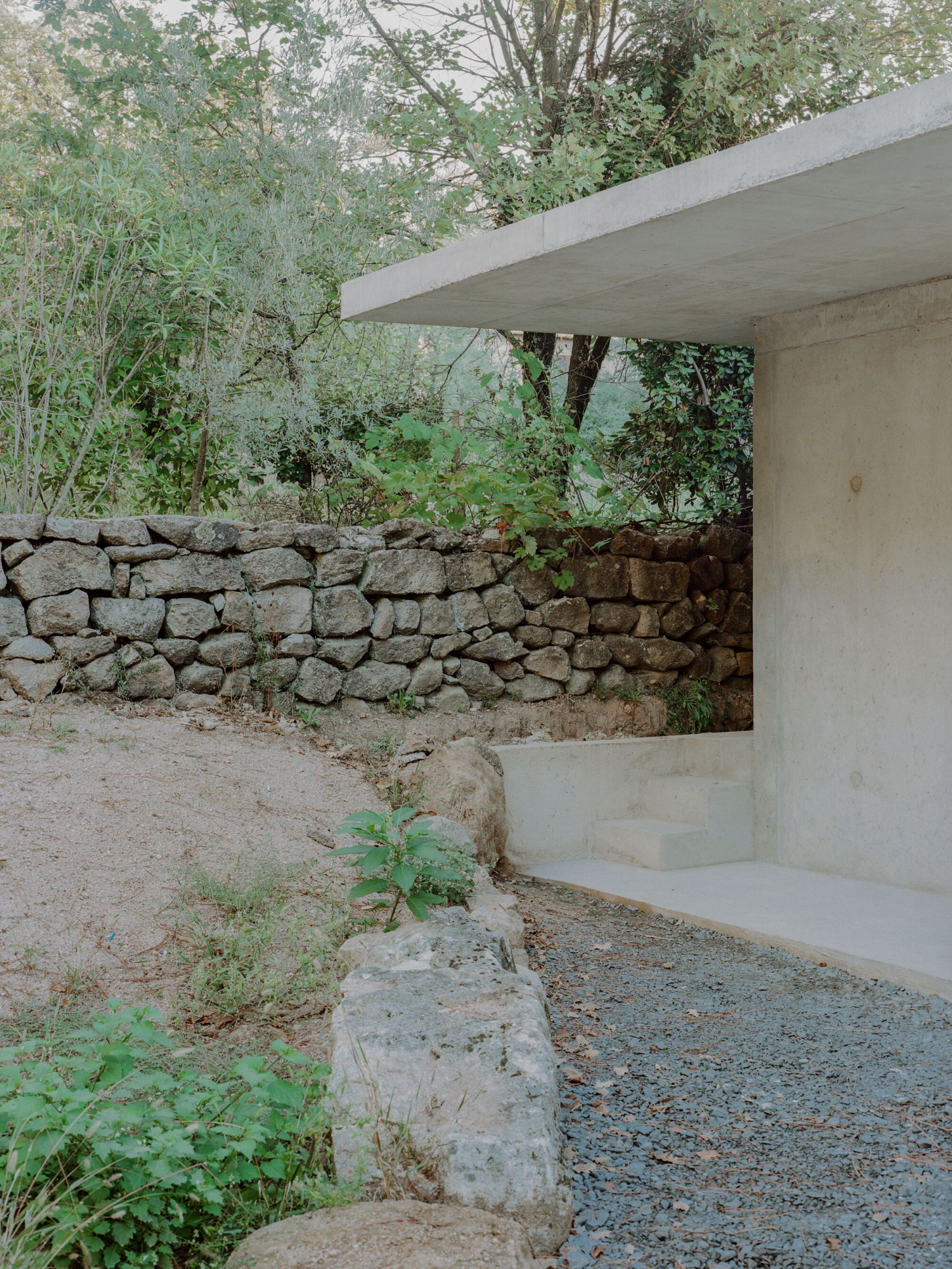 PAYZ House by Alors Studio — A concrete cantilevered shelter extends over a gravel courtyard, its monolithic slab creating a sharp horizontal line against dense vegetation.