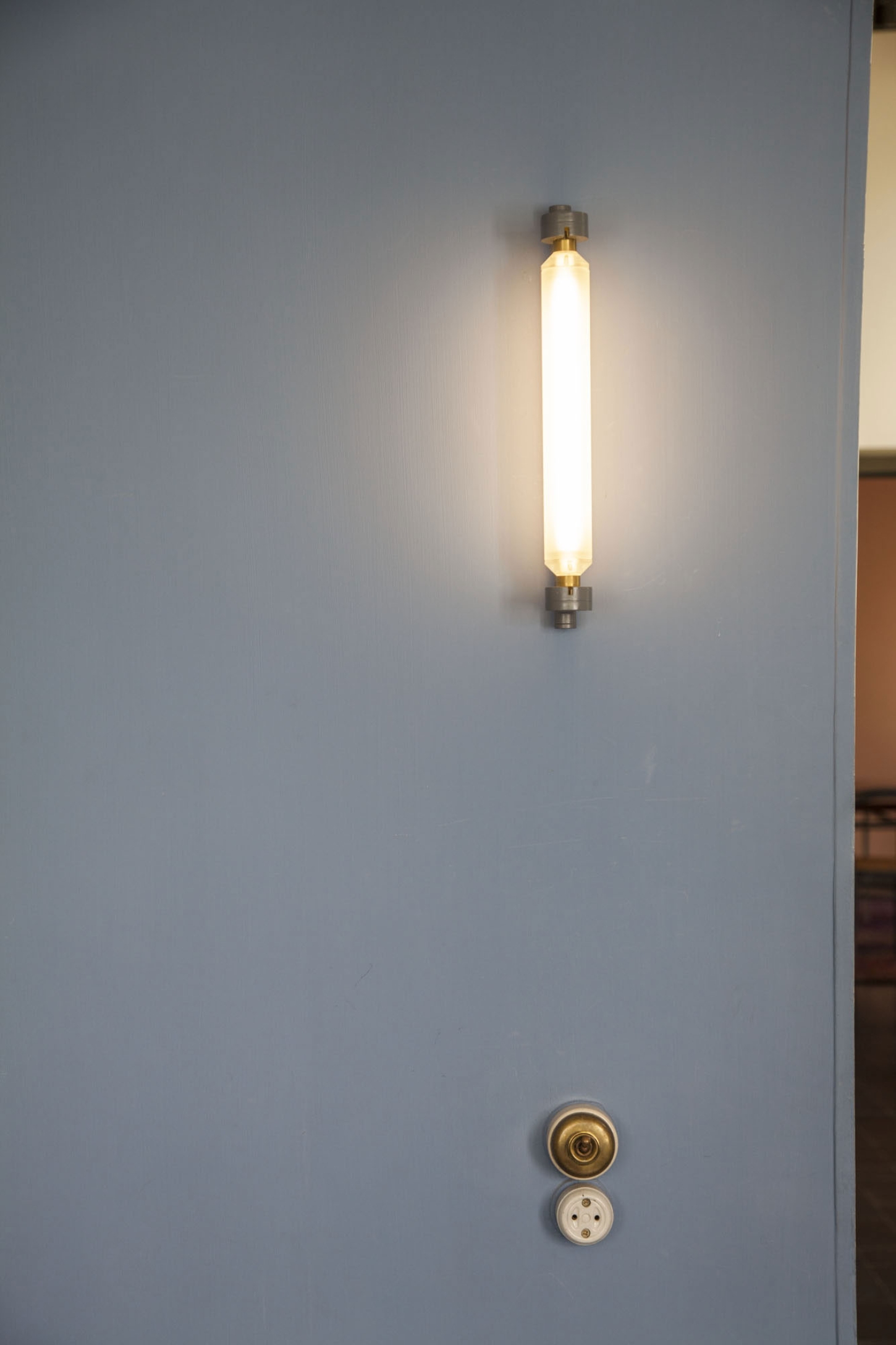 Maison La Roche-Jeanneret by Le Corbusier by Le Corbusier and Pierre Jeanneret — A vertical tubular brass or bronze fixture mounted centrally on white plaster wall, emitting warm fluorescent light through frosted glass cylinder.