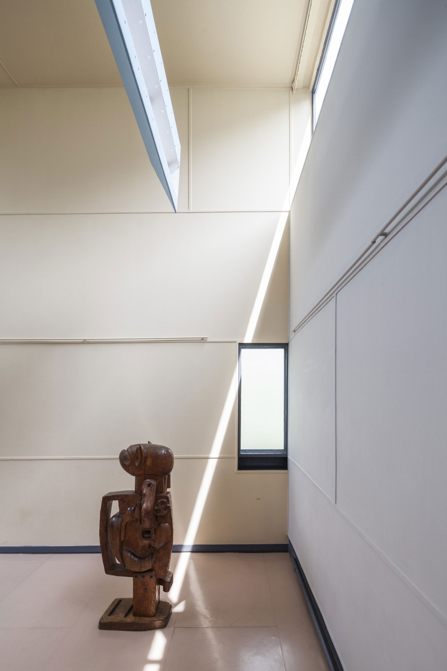 Maison La Roche-Jeanneret by Le Corbusier by Le Corbusier and Pierre Jeanneret — A wooden sculptural figure occupies the center of a gallery corridor, its carved form casting sharp shadows across polished concrete flooring.